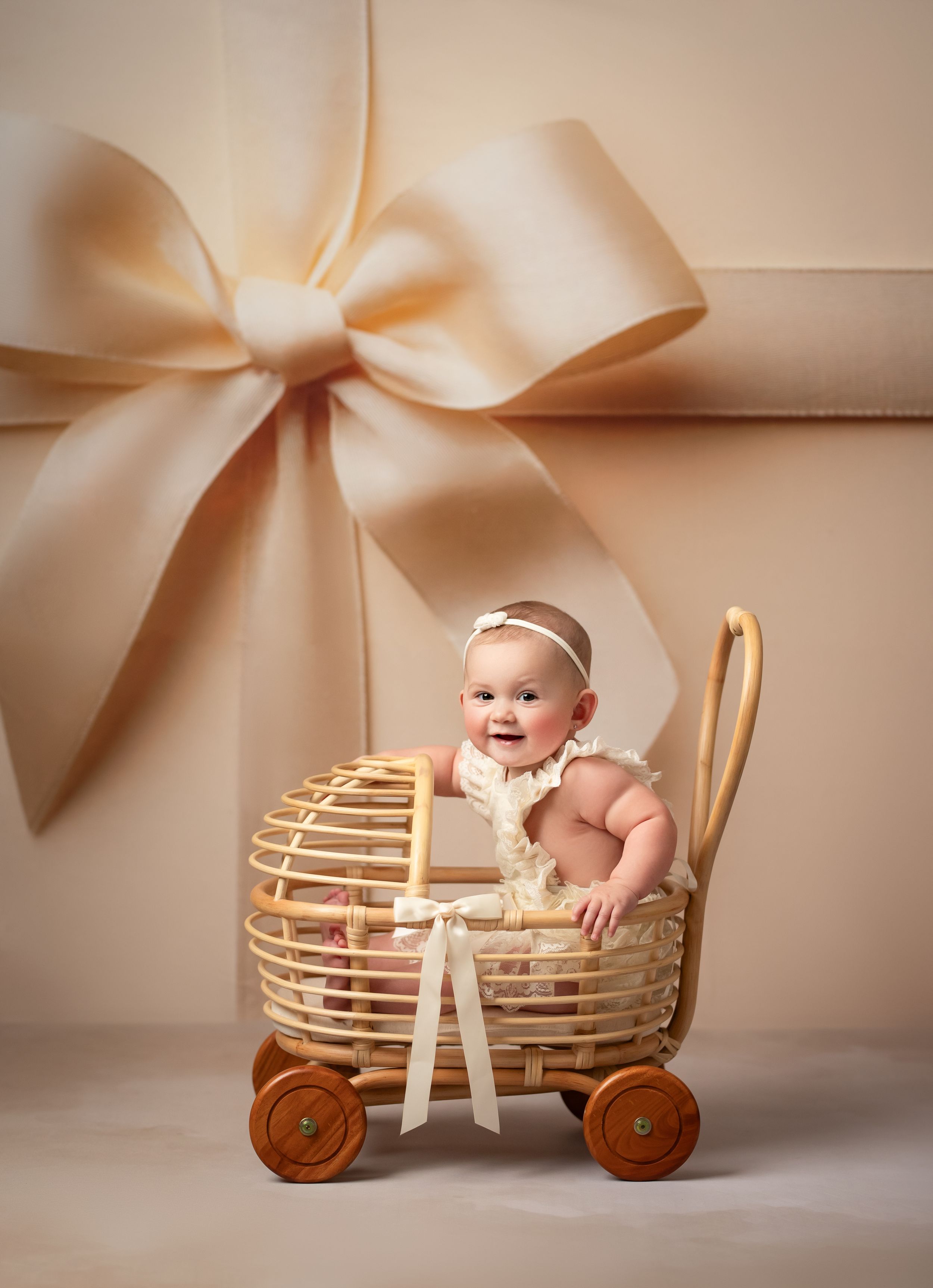 A baby sits in a decorative wooden wagon prop against a beige backdrop with a large bow accent.