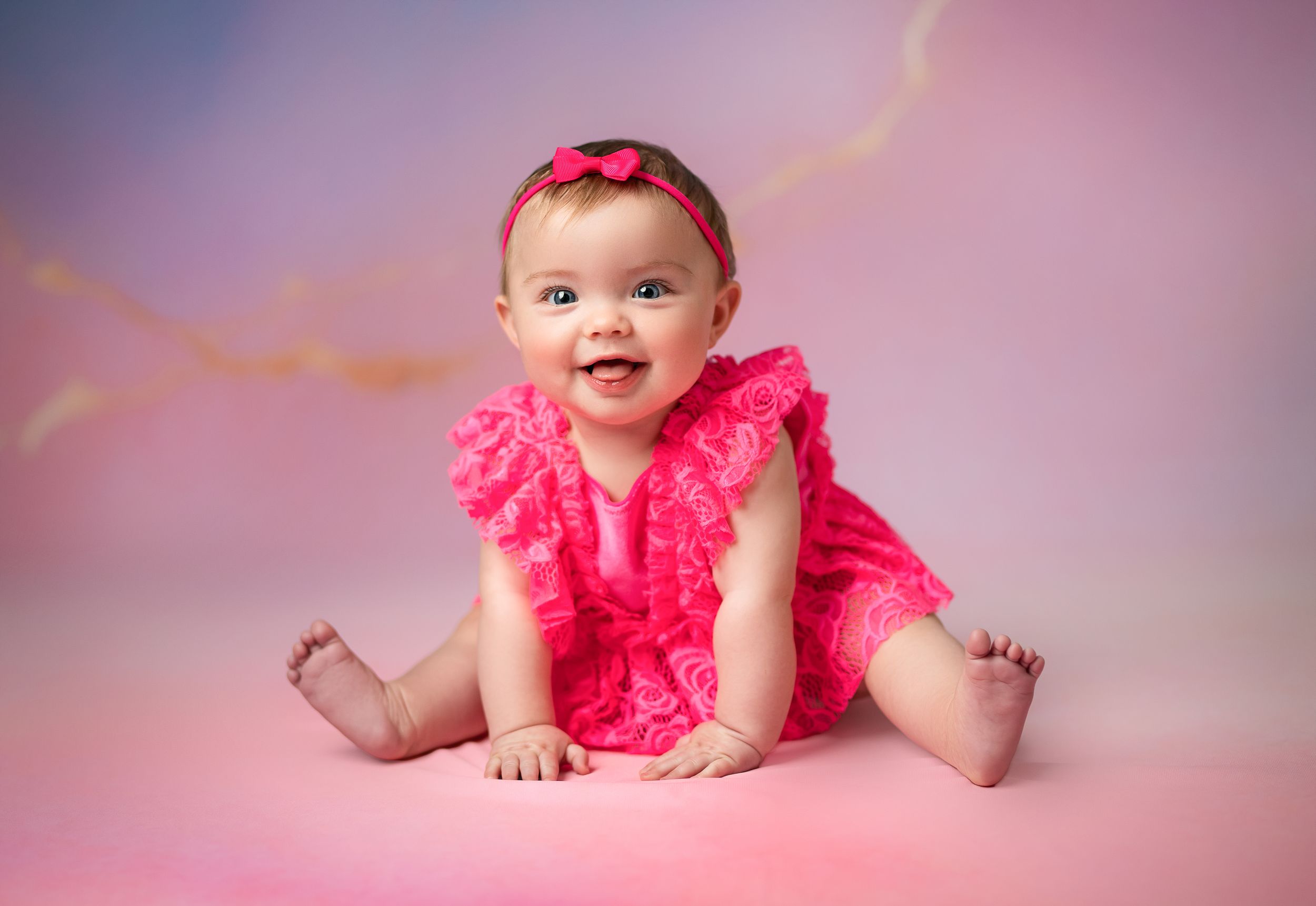 A baby wearing a bright coral pink ruffle dress poses on a pink backdrop during a professional photo session.