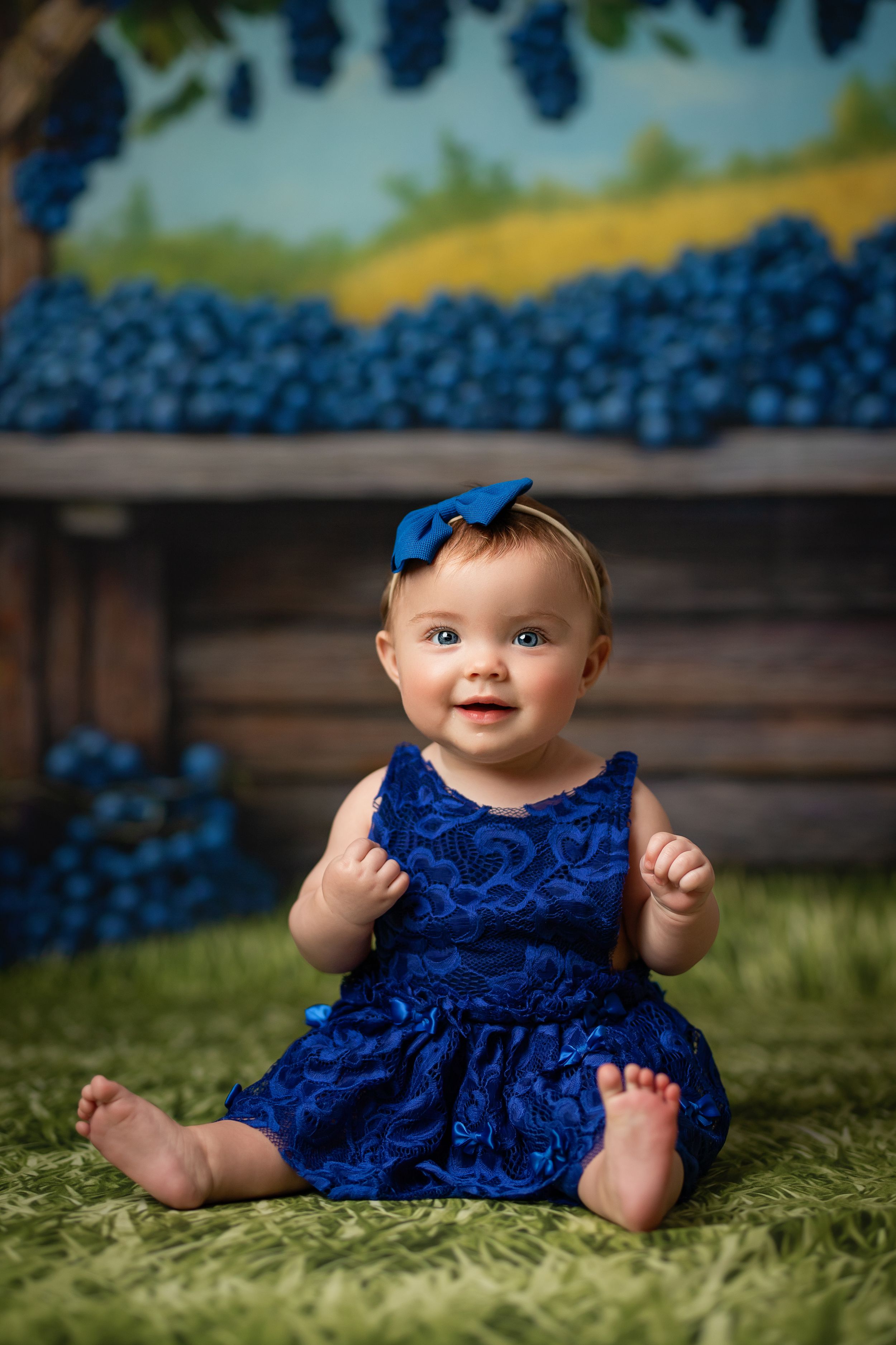 A baby in a navy blue dress and matching bow sits on green grass with blue flowers in the background.