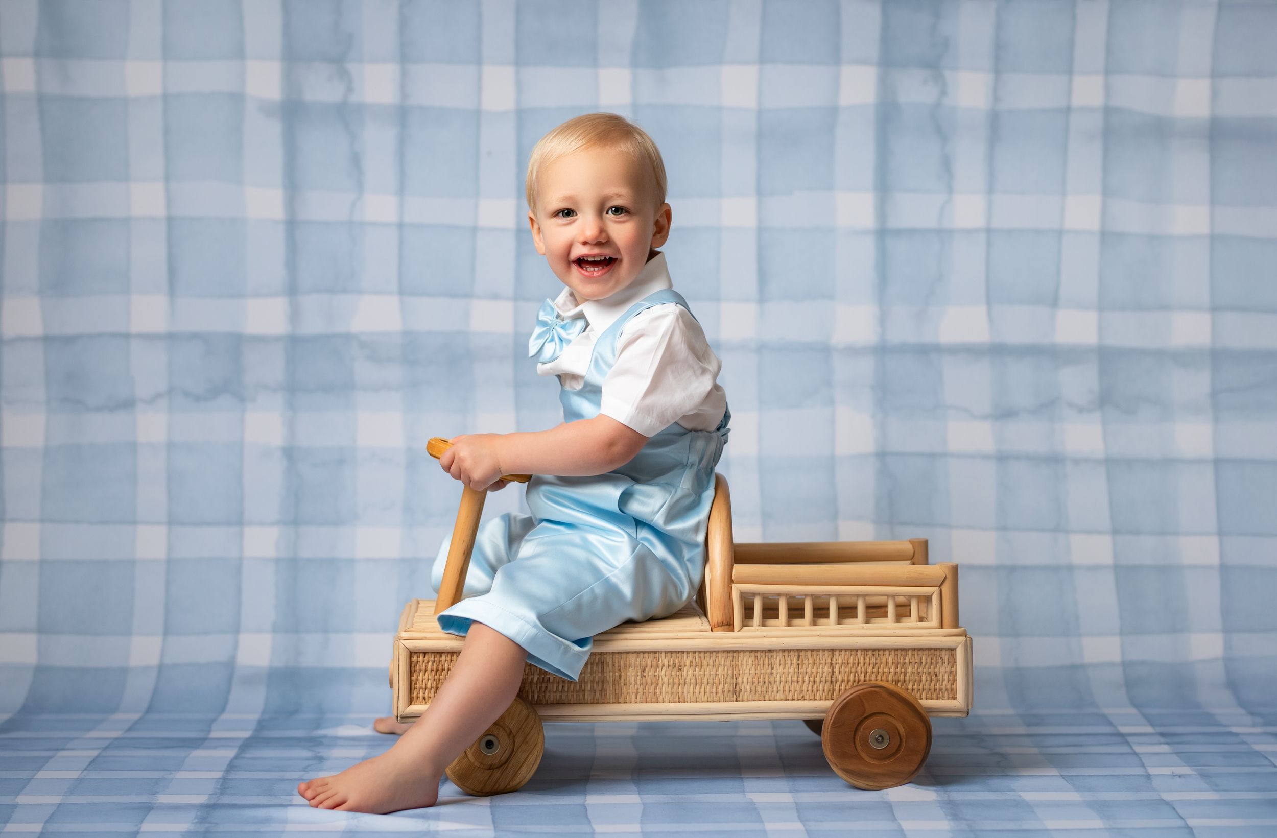 A toddler in blue and white outfit sits on a wooden toy wagon against a blue checkered backdrop.