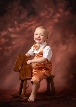 Portrait of a cheerful baby in orange overalls and bow tie sitting on a wooden rocking horse against a dark backdrop.