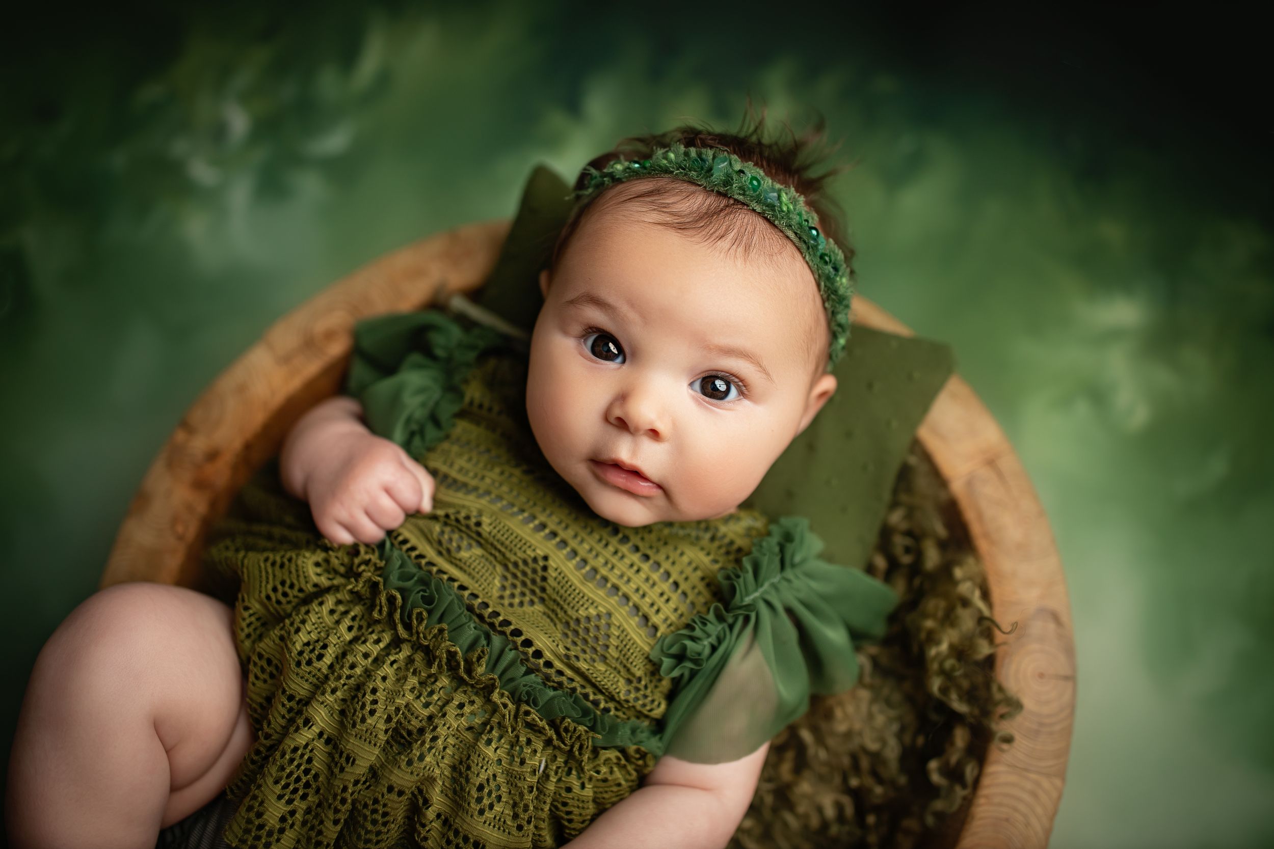 Newborn baby wearing green knit outfit lying in wooden bowl against forest green backdrop.