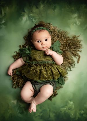 Baby wearing olive green knit dress lies on green backdrop in artistic newborn photography series.
