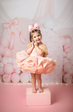 Little one in a ruffled peach dress and bow stands on pink box against pastel balloon backdrop.