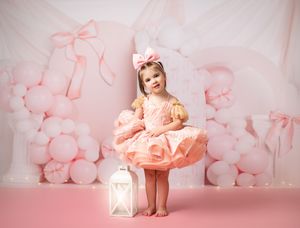 Little girl in peach tutu dress poses with lantern against pink balloon backdrop and bows for birthday photoshoot.