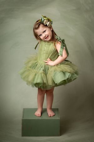 A small dancer in a green tulle dress stands barefoot on a green box against a soft sage-colored backdrop.