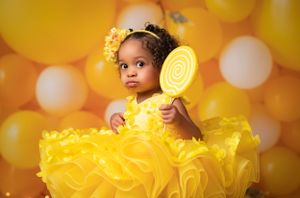 Child in bright yellow tulle dress poses with lollipop props against orange bokeh background.