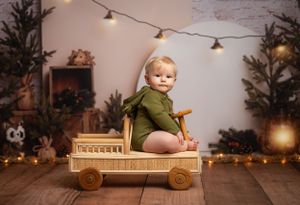 Toddler in green bodysuit sits on wooden toy wagon with string lights and holiday decor in background.