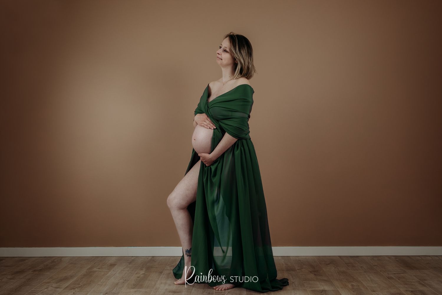 Zwangerschapsfoto van een vrouw in een studio, gekleed in een elegante, vloeiende scarf. Ze is ontspannen met haar handen op haar buik.