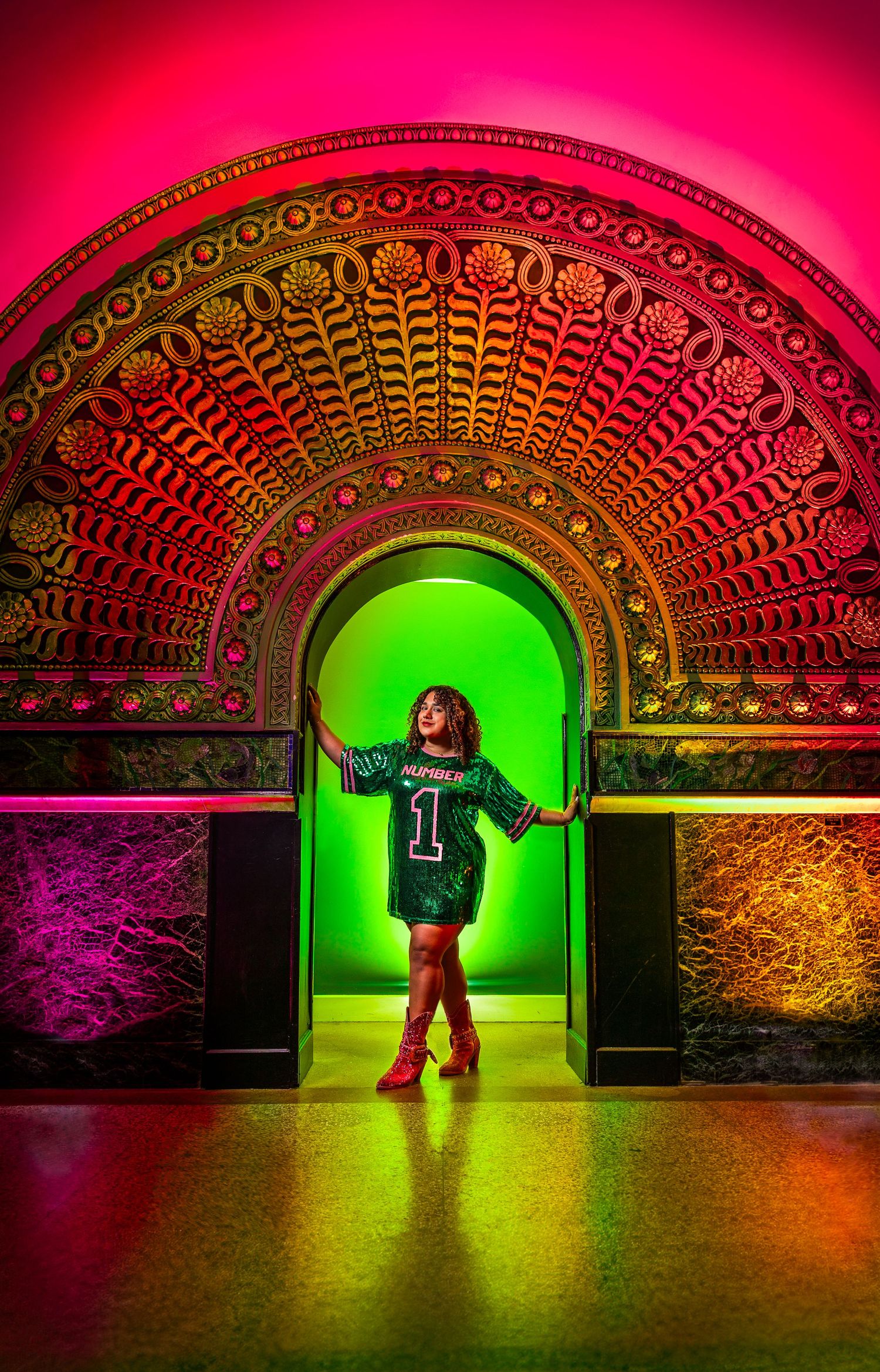 Carolina Waldroff in a sparkling green jersey dress poses confidently under an ornate archway lit with vibrant rainbow lighting. STL, MO