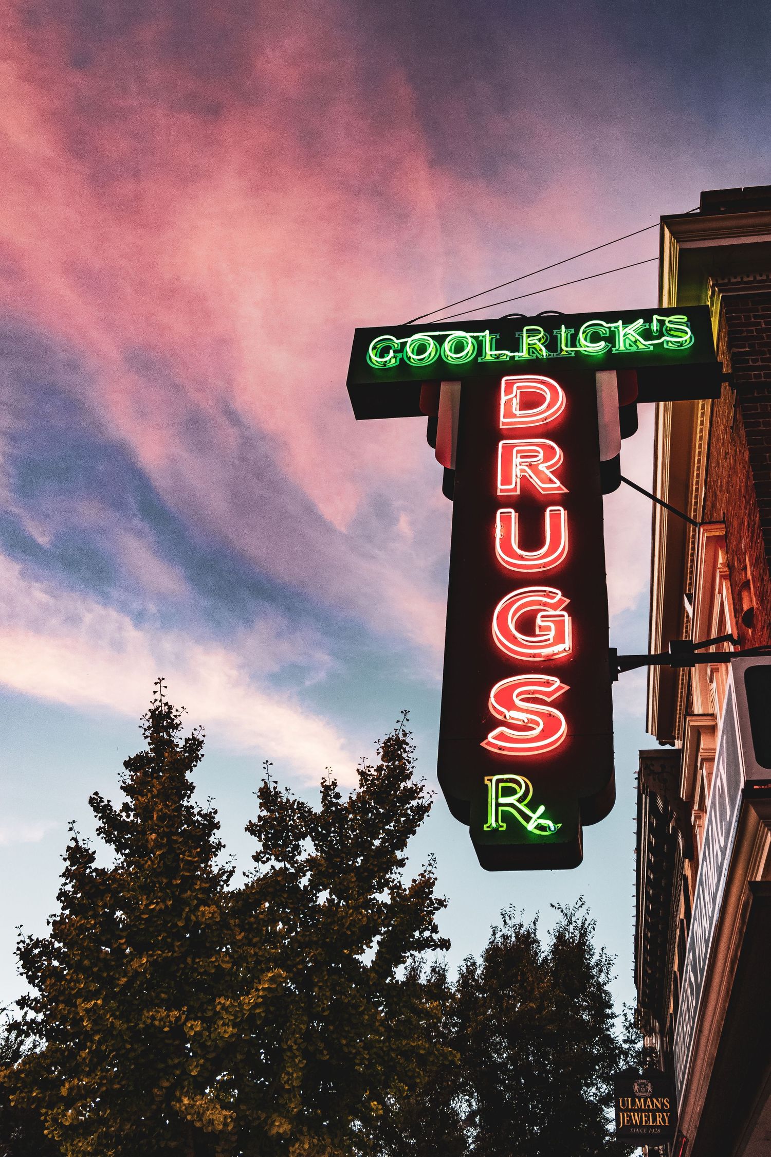 Neon sign letters spelling 'DRUG STORE' illuminate against a pink and purple sunset sky with silhouetted trees.