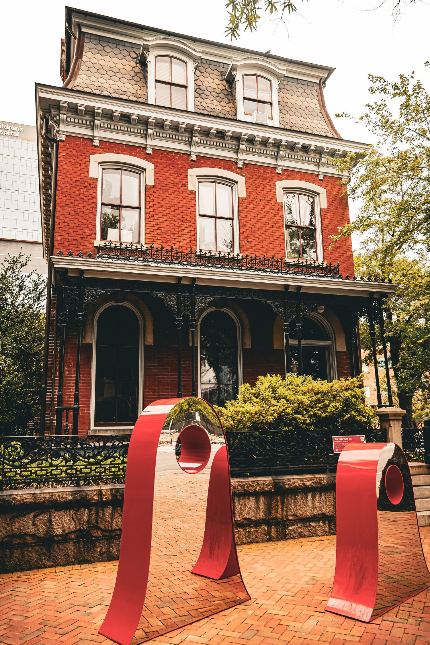 Red curved art sculptures line a brick walkway in front of historic Victorian-style buildings with ornate details.