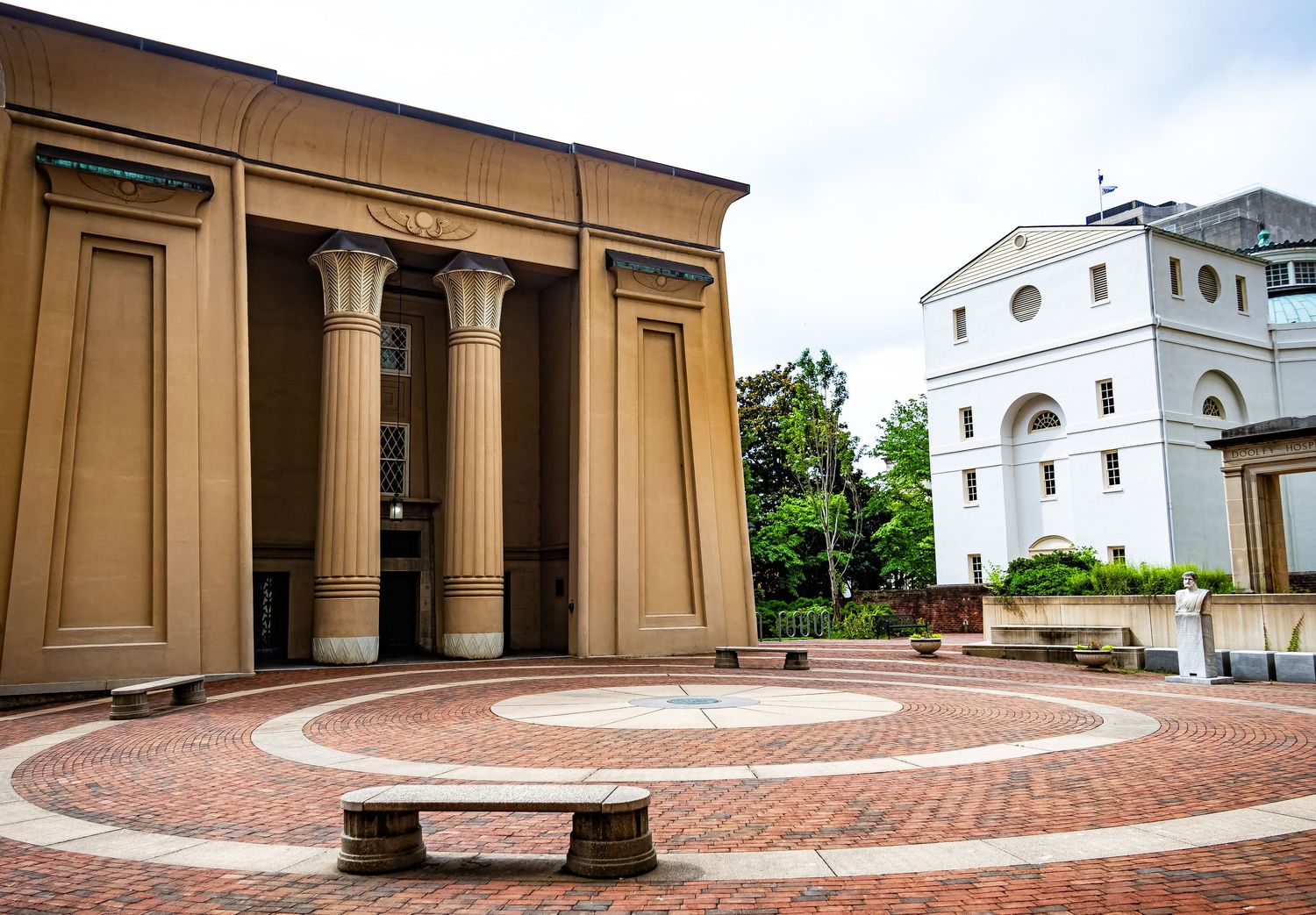 Classical columned building entrance with brick courtyard and curved stone bench near white church architecture.
