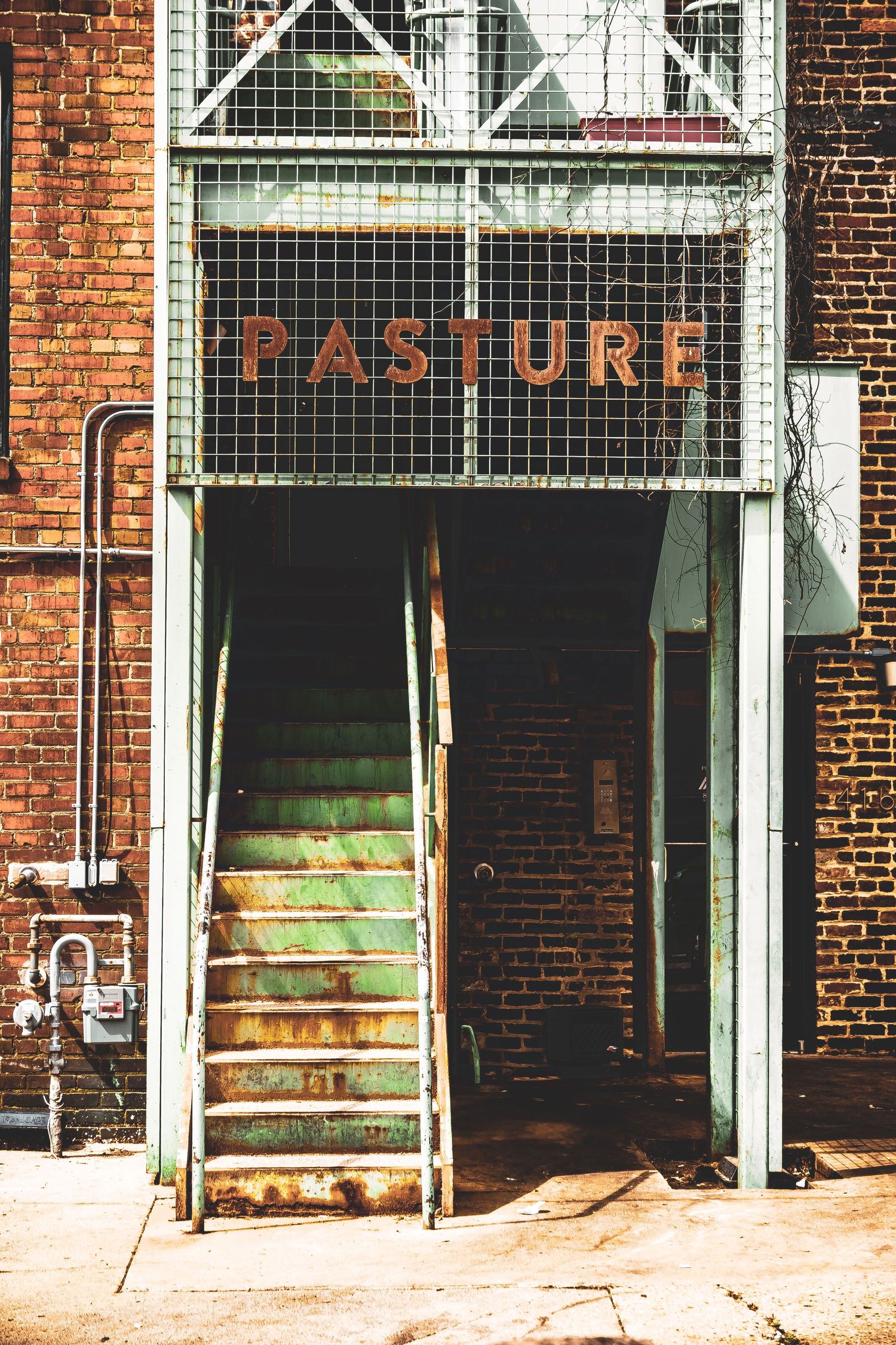 Rustic brick building entrance with weathered metal stairs and 'PASTURE' signage above a gated doorway.