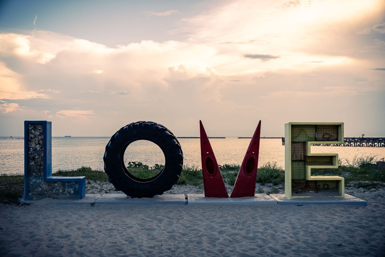 Large colorful letters spell out LOVE against a sunset sky on a beach with ocean waves in the background.