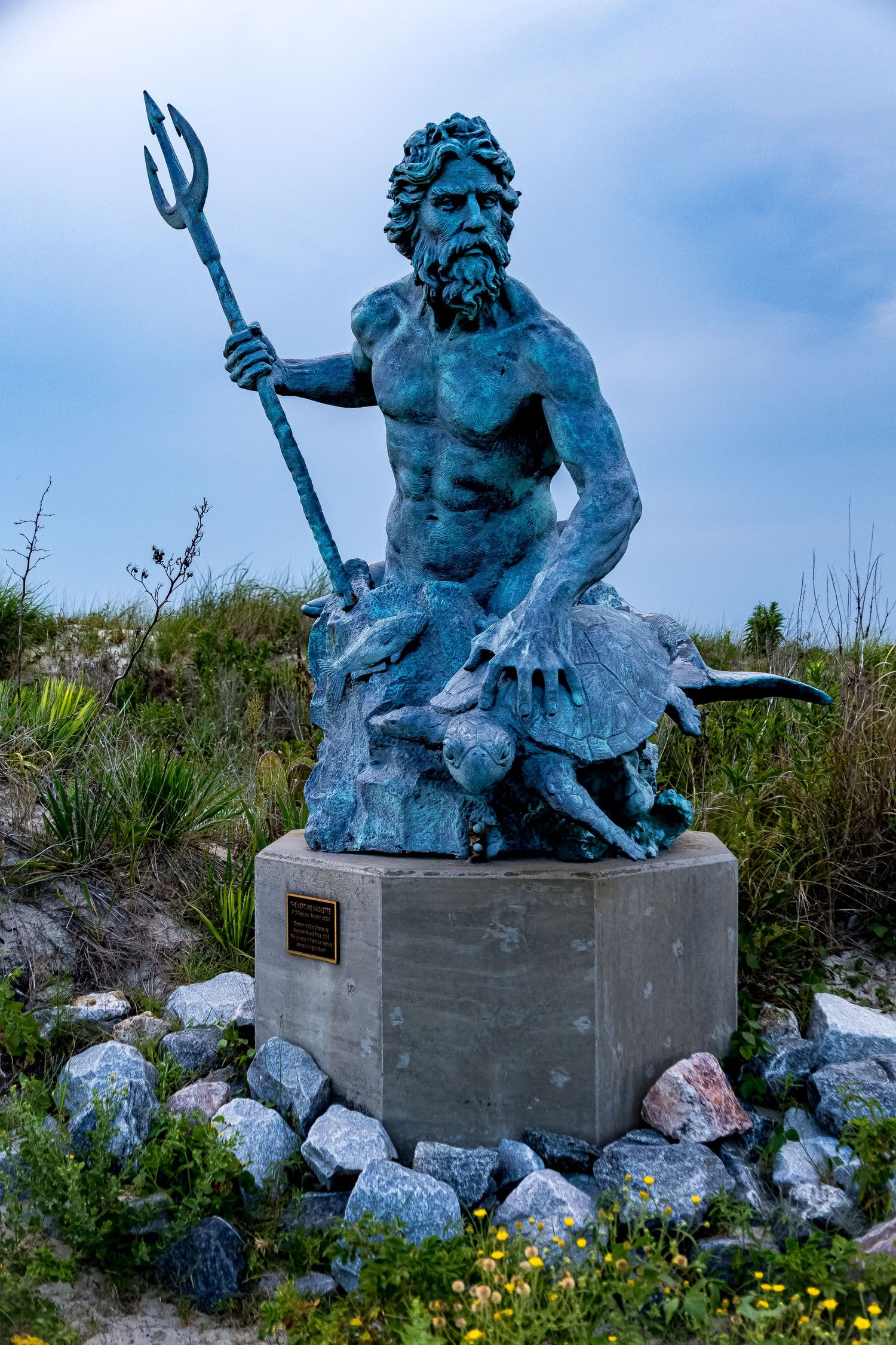 A large blue-tinted statue of King Neptune holding a trident sits on a concrete base surrounded by rocks and wildflowers.