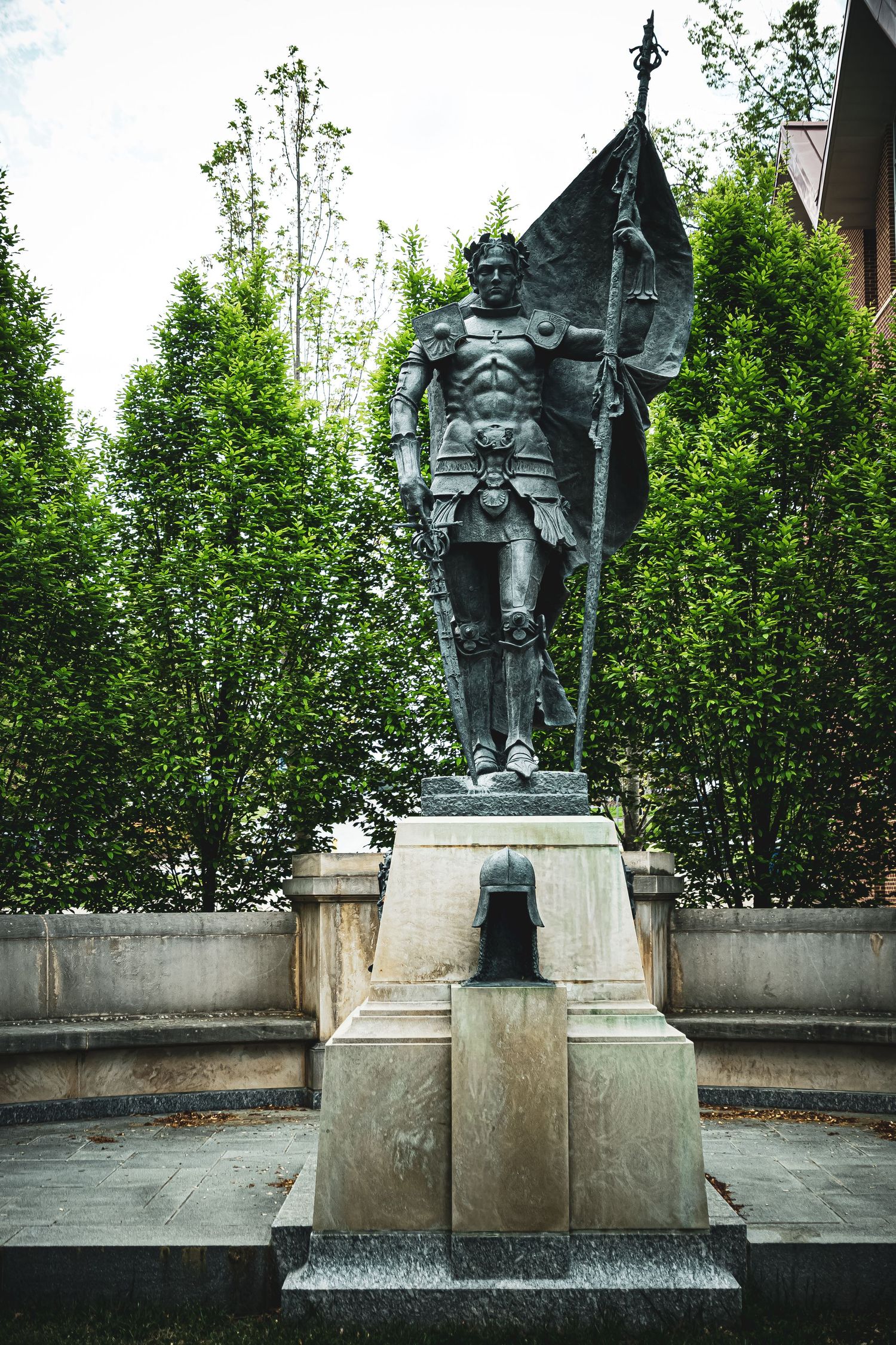 A series of four identical bronze statues on stone pedestals surrounded by green trees and foliage.