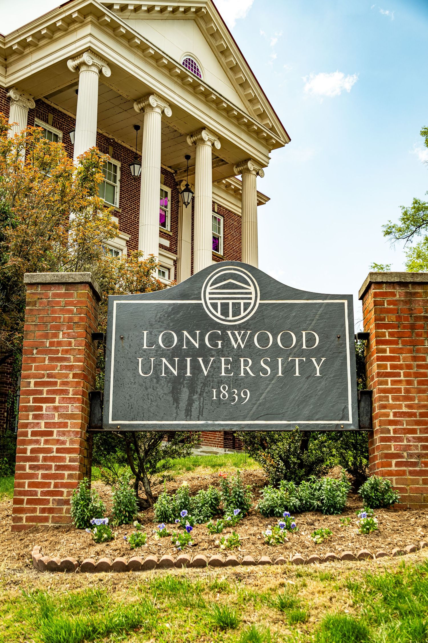 Three identical stone entrance signs for Longwood University mounted on brick walls with landscaping and columns.
