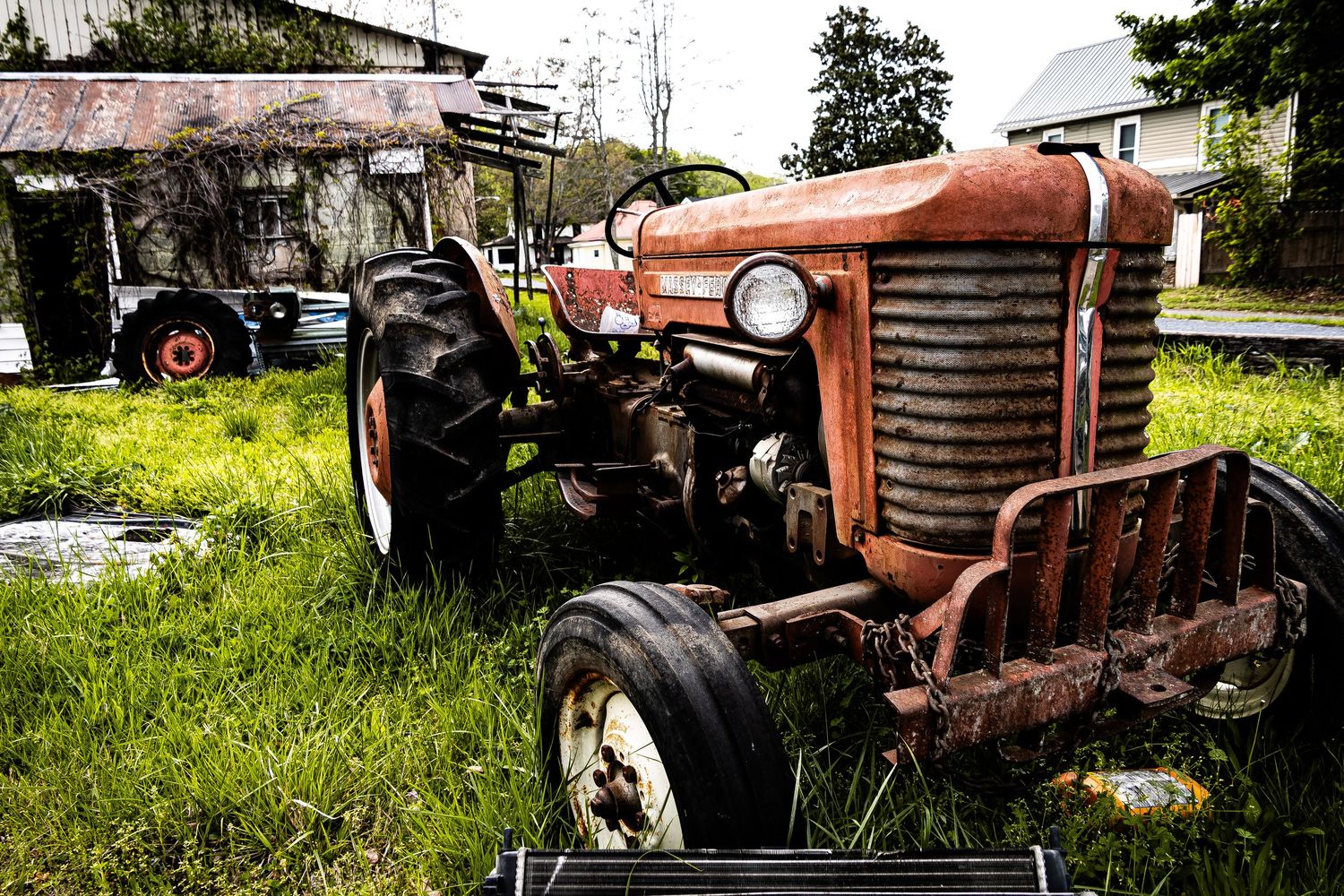 Old rusty red Massey Ferguson tractor sits abandoned in overgrown grass near a farm building.