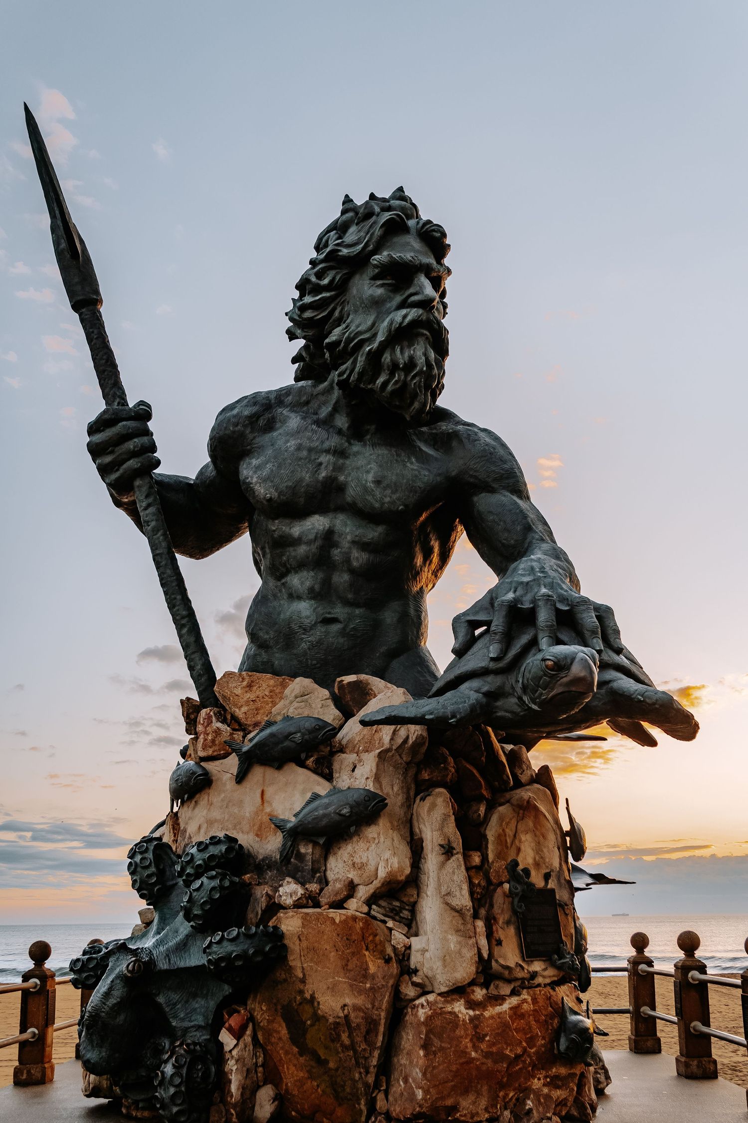 Large bronze statue of King Neptune holding a trident stands against a sunset sky at Virginia Beach oceanfront.