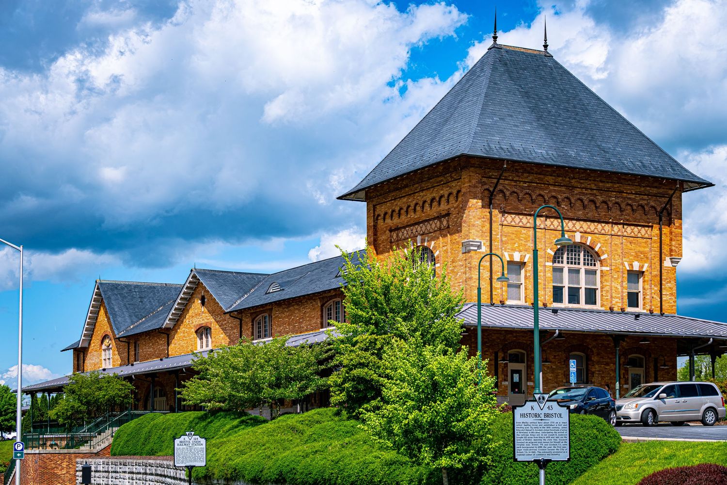 A panoramic view of a historic brick building with a distinctive tower and arched windows against a cloudy blue sky.