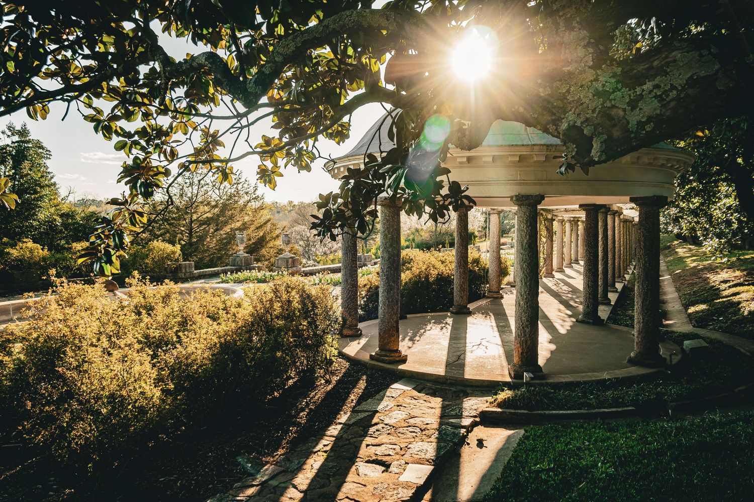 Sun rays shine through trees and gazebo creating a golden silhouette against a serene waterfront setting.