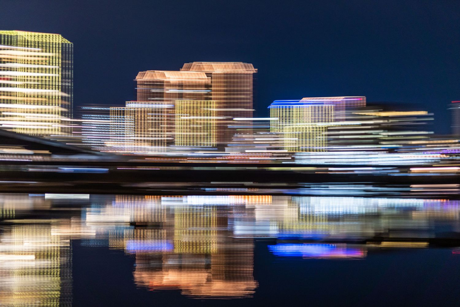 Abstract cityscape with motion blur effect showing illuminated buildings reflecting in water at night.
