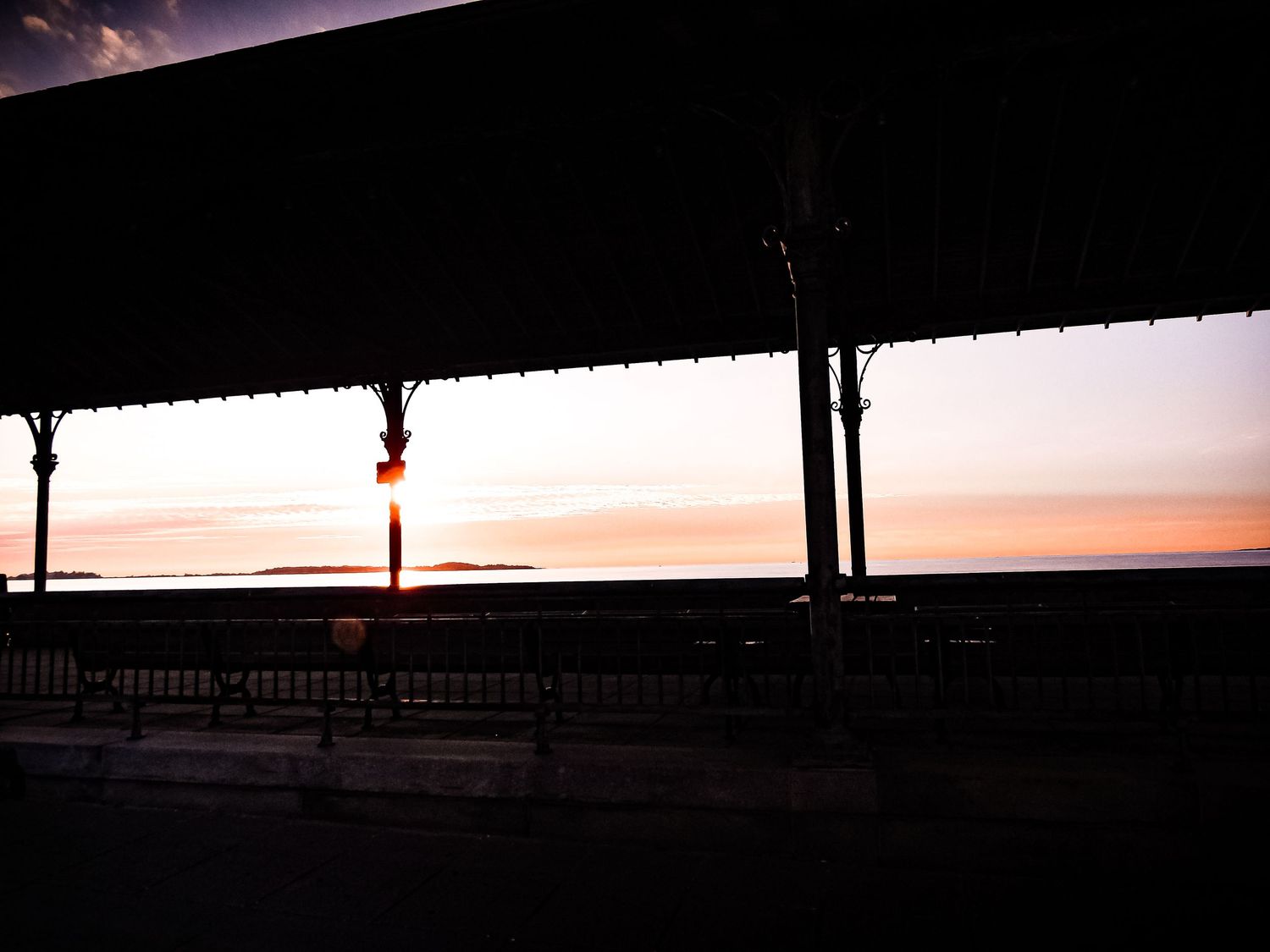 A panoramic view of a sunset silhouetting a bridge and waterfront structures against an orange and purple sky.