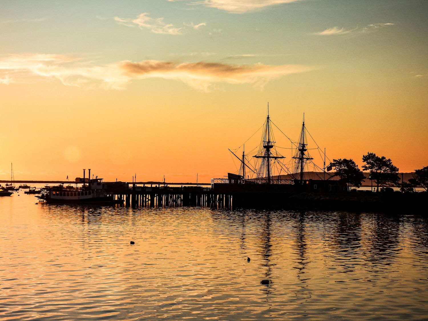 A panoramic view of a harbor at sunset with silhouettes of boats and docks against an orange and pink sky.