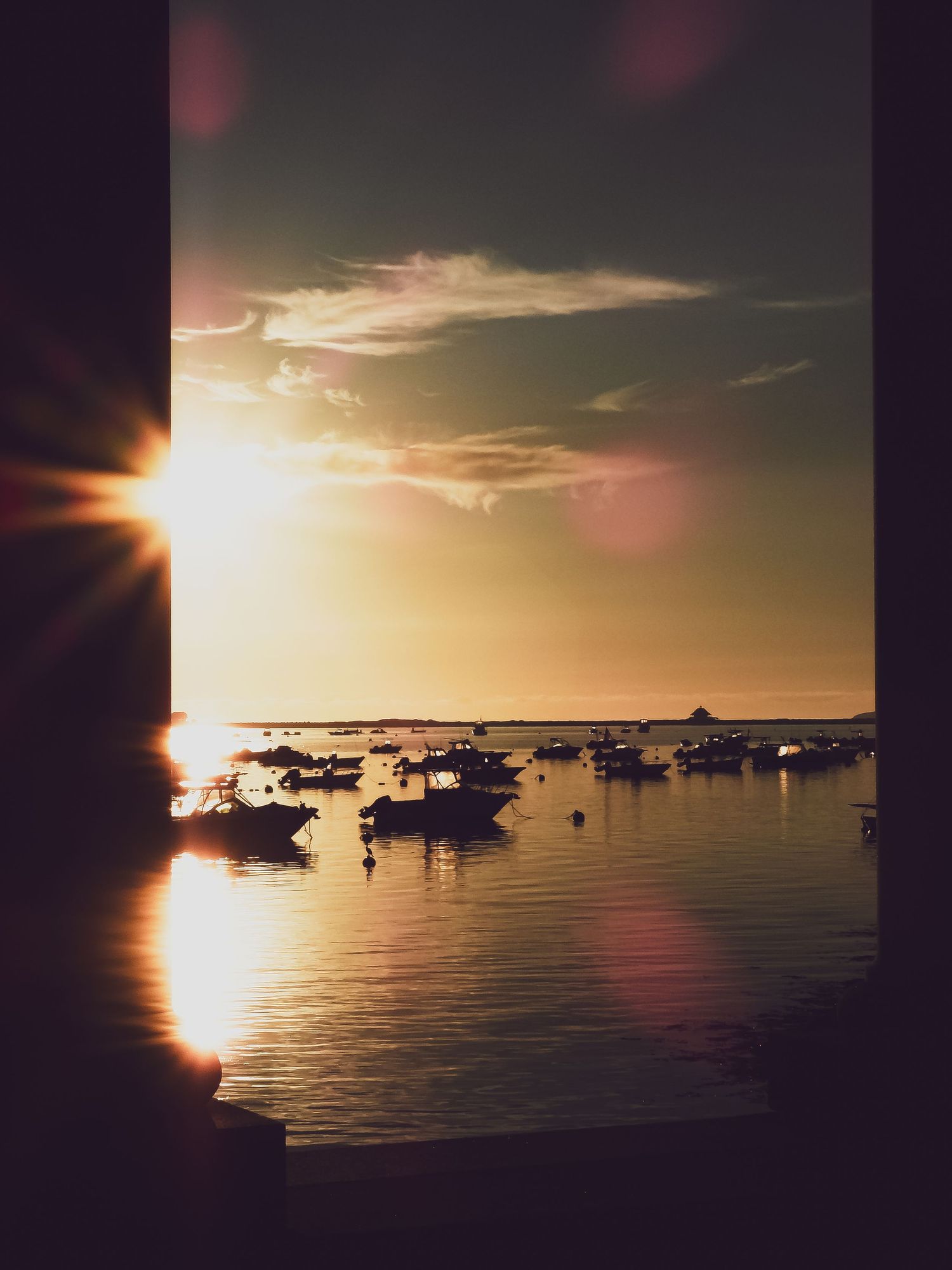 Boats silhouetted against a golden sunset over calm harbor waters with lens flare and warm tones.