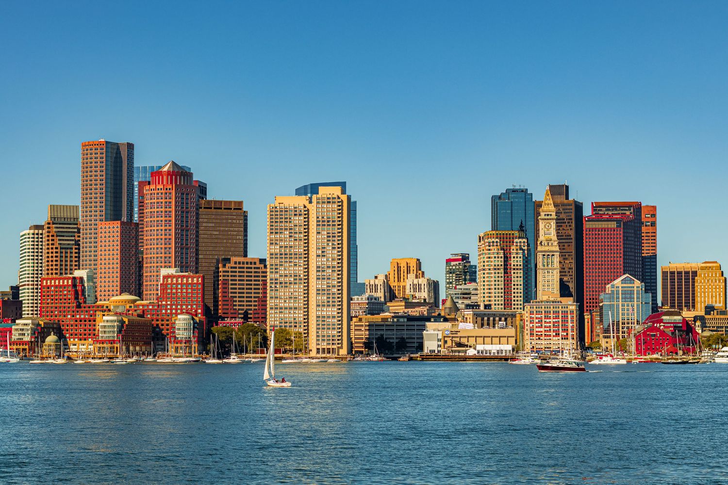 Boston skyline with waterfront buildings and sailboat on harbor during sunny day against clear blue sky.