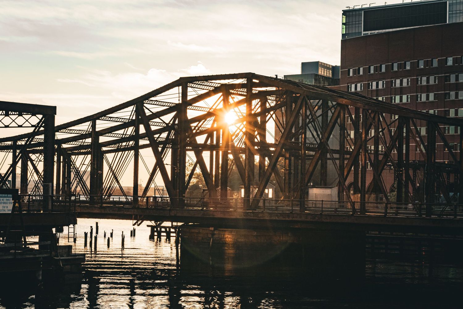 Sunlight peeks through an industrial steel bridge over calm water at sunset with city buildings in the background.