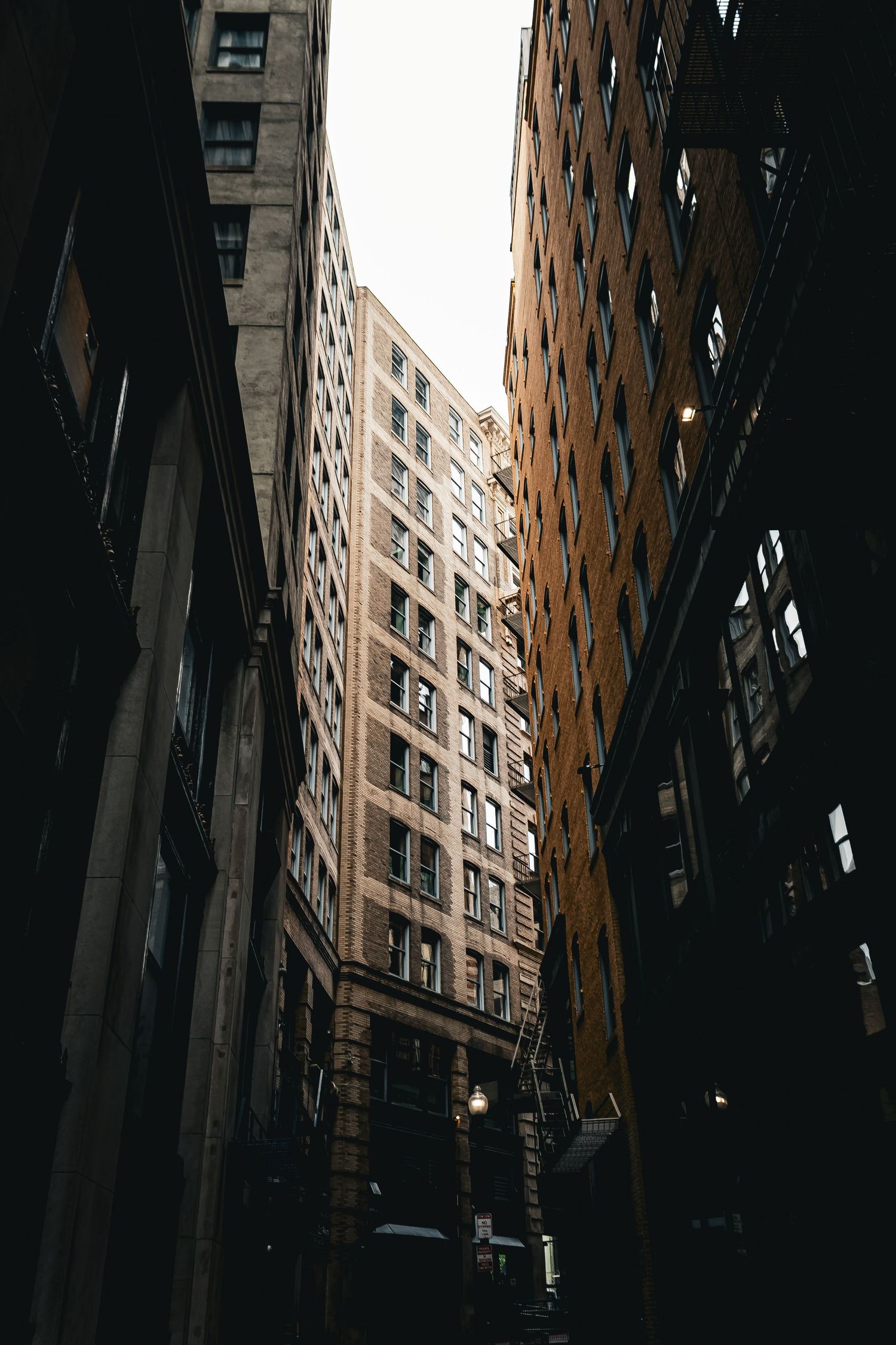 Looking up at tall brick apartment buildings in a narrow urban alley with dramatic shadows and lighting.
