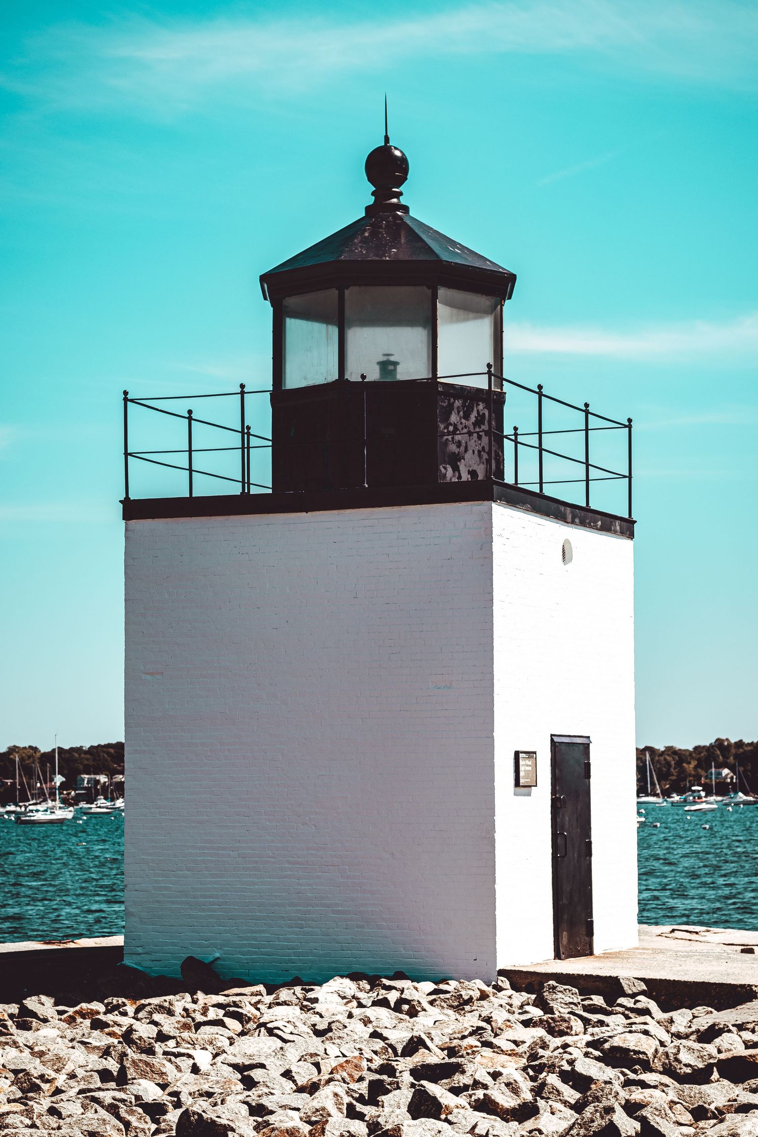 A small white lighthouse with a black top sits on a rocky shoreline against a bright turquoise ocean and sky.