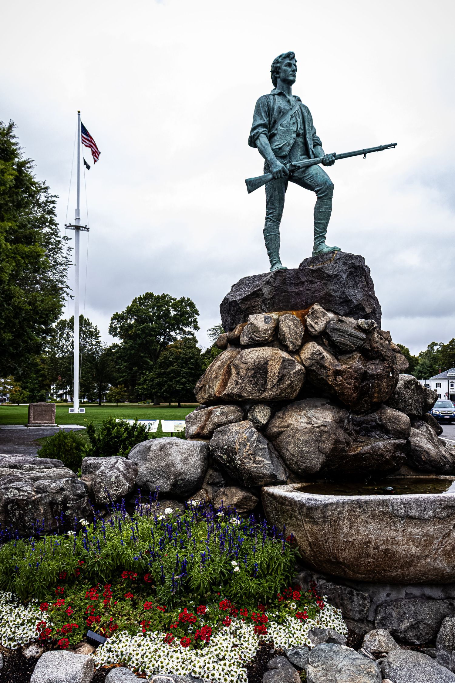 Bronze Minuteman statue stands atop stone monument with American flag and colorful flower garden at base.