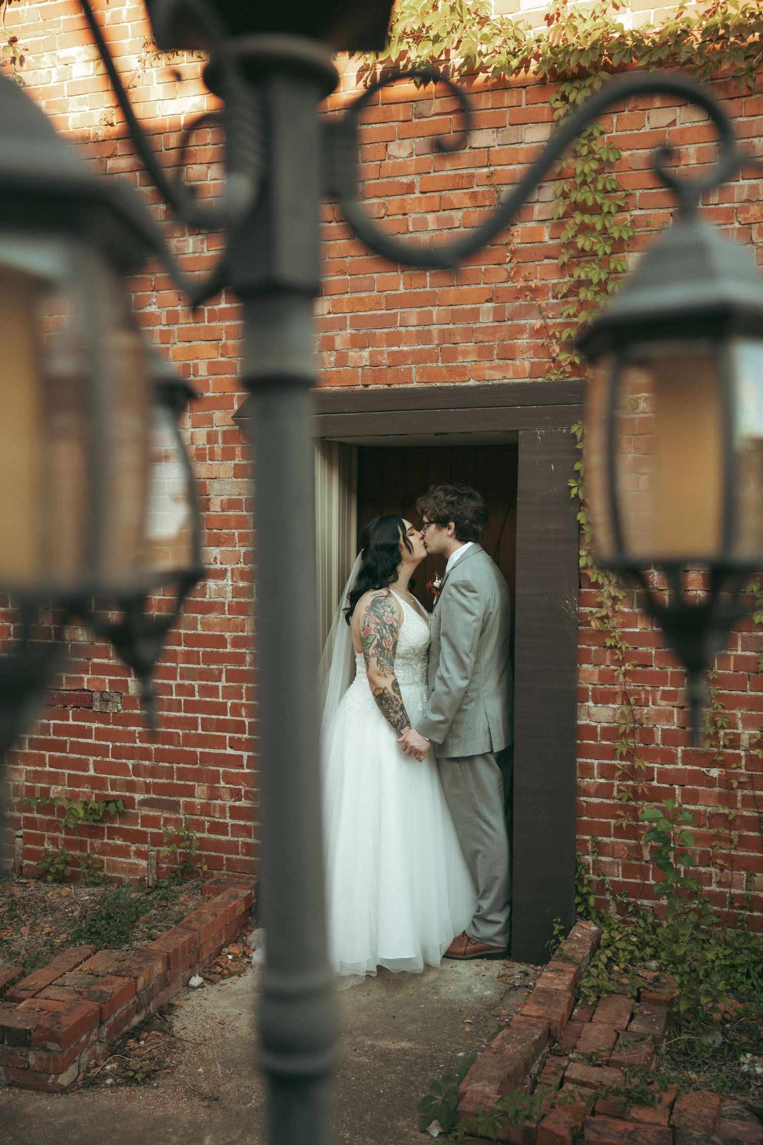 A romantic moment captured through vintage lanterns as a couple embraces in a brick doorway wearing wedding attire.