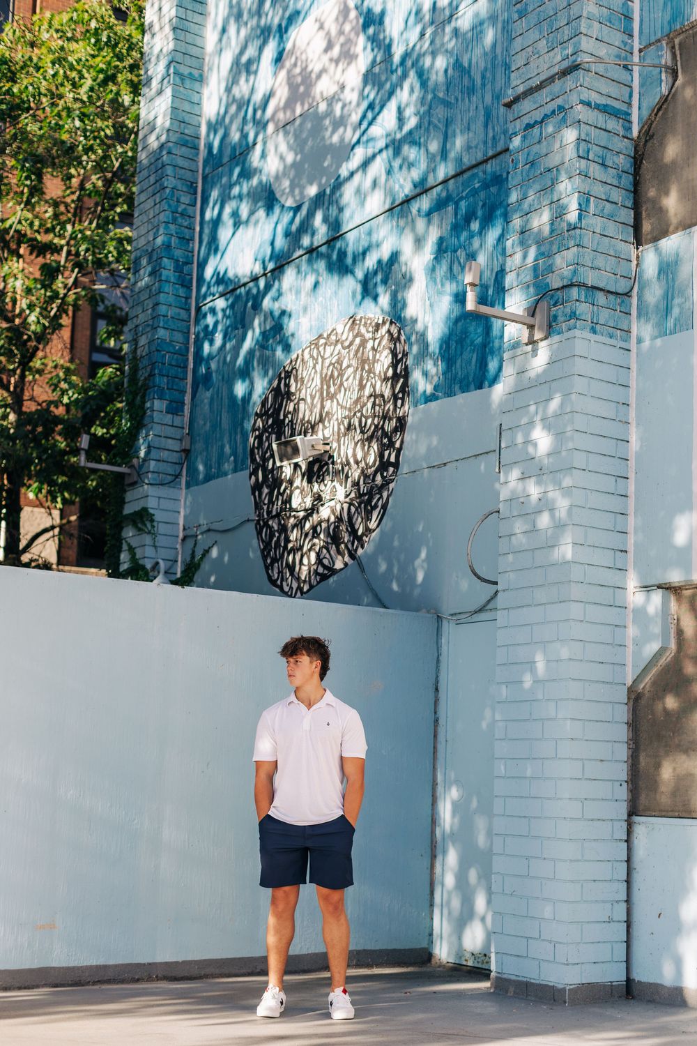 Person in white t-shirt and navy shorts poses against modern blue brick wall with sunlight casting artistic shadows.