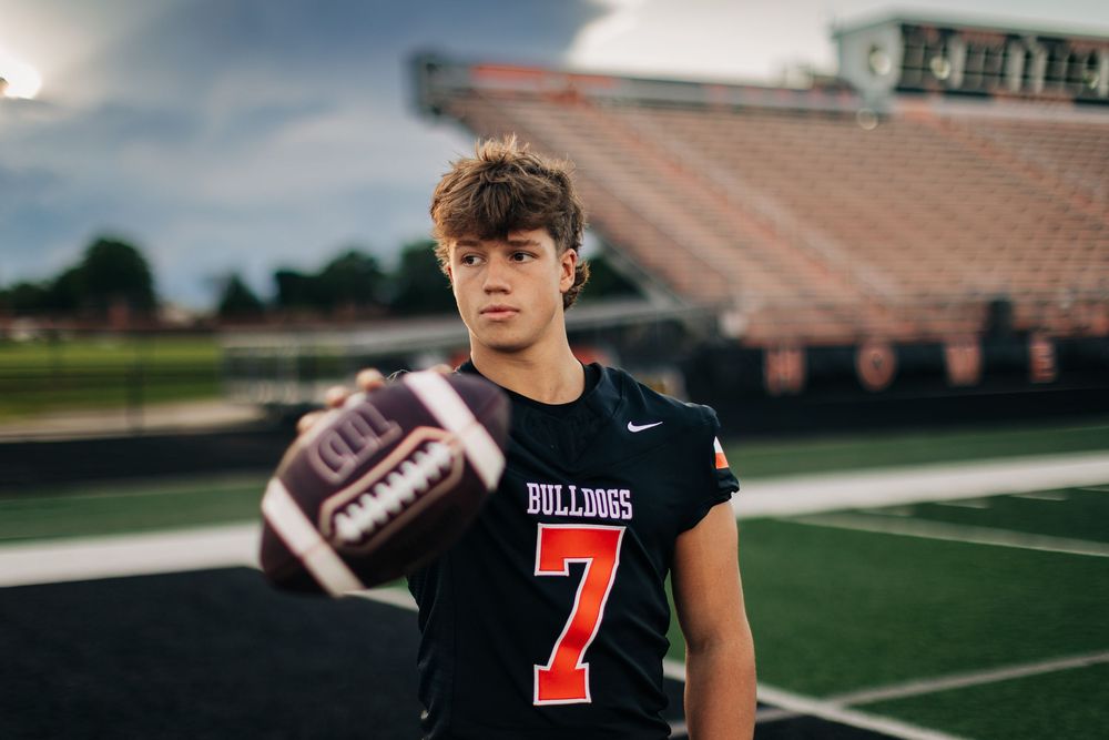 Football player in jersey number 7 stands on the field during dusk with stadium lights glowing in background.