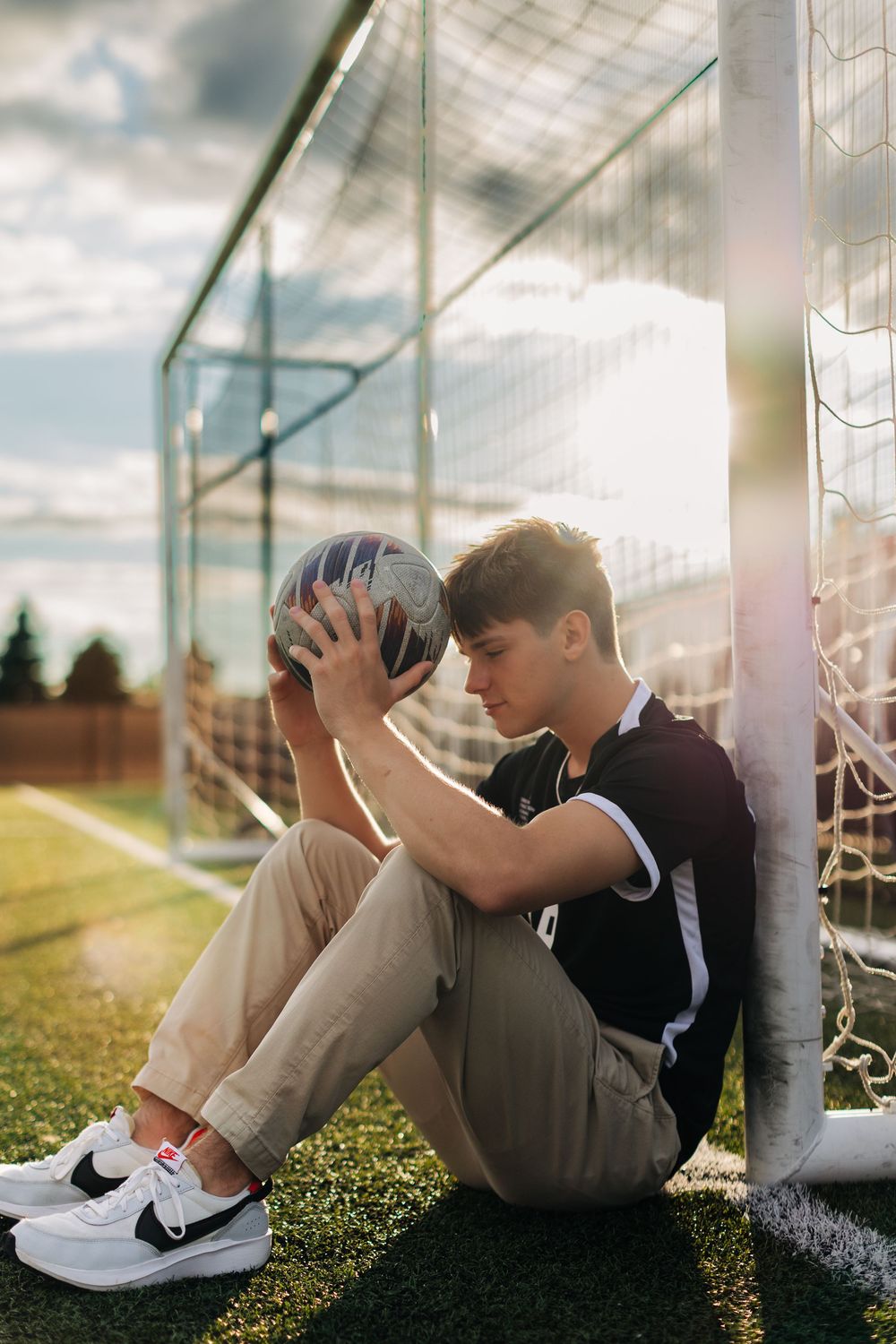Person sitting by soccer goal post on grass field during sunset with dramatic lighting and shallow depth of field.