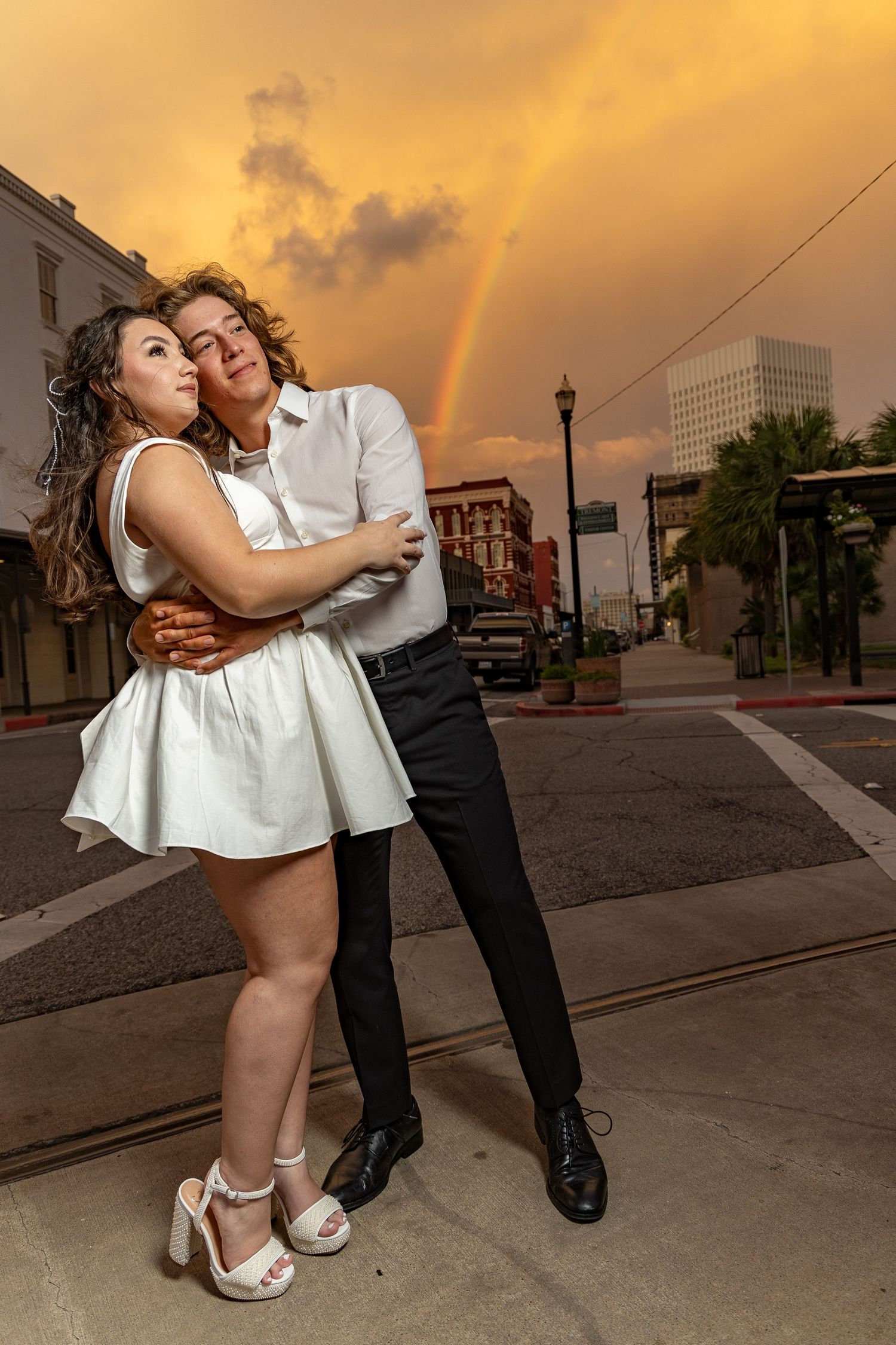 Couple in formal wear posing at sunset on city street with golden sky.