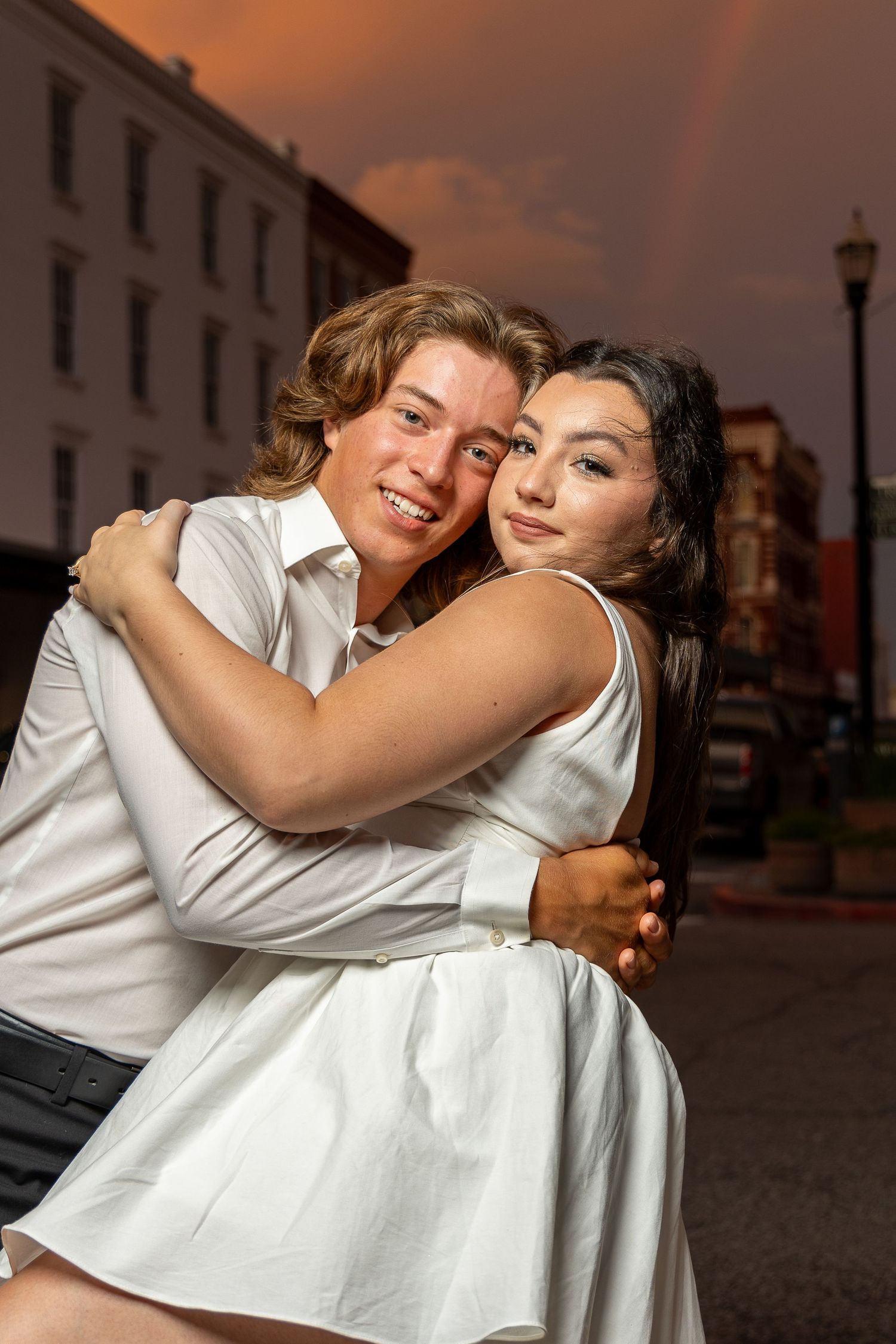 A romantic couple in white outfits embrace and smile during a sunset photoshoot on a city street.