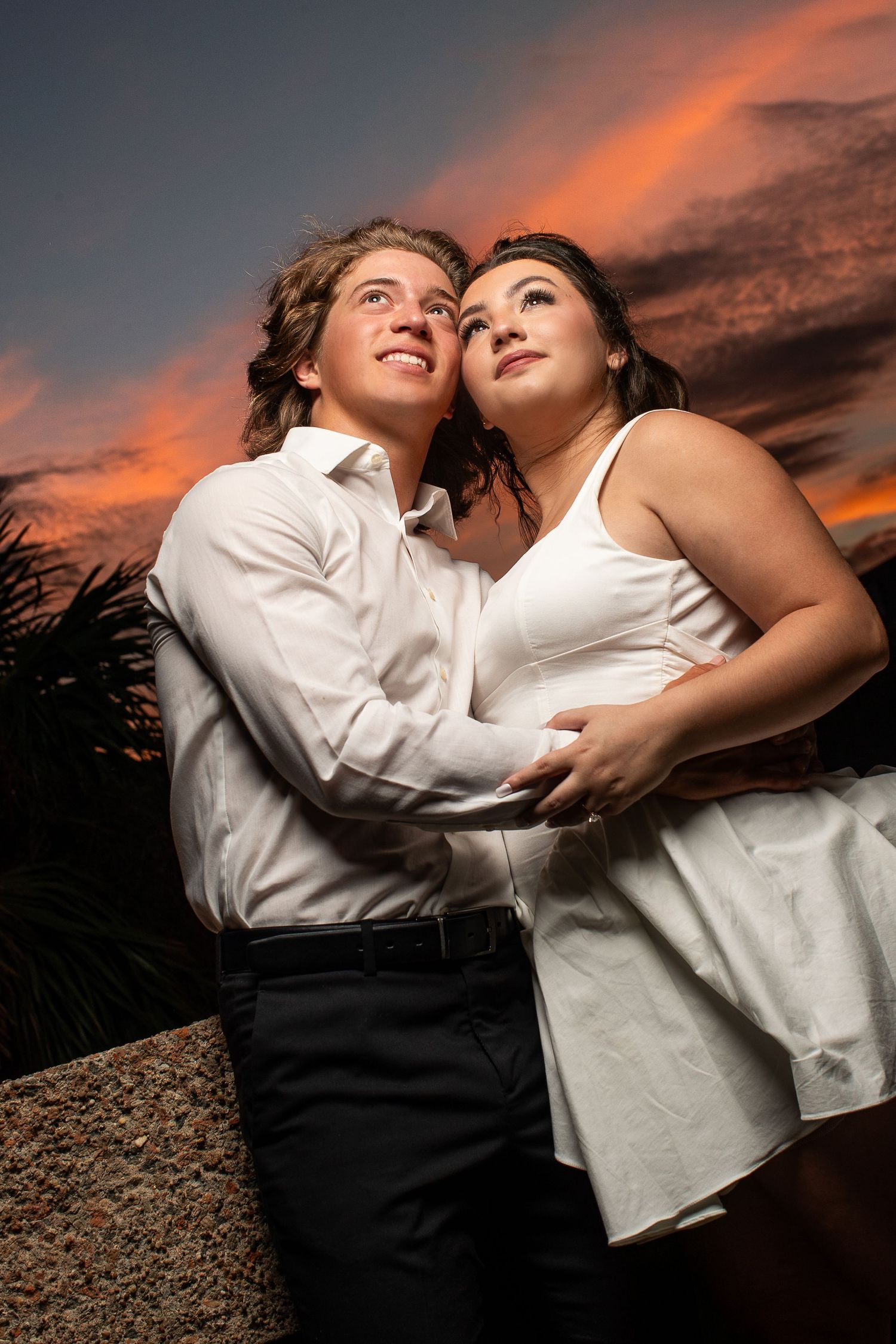 A couple in white clothing poses together against a dramatic orange sunset sky.