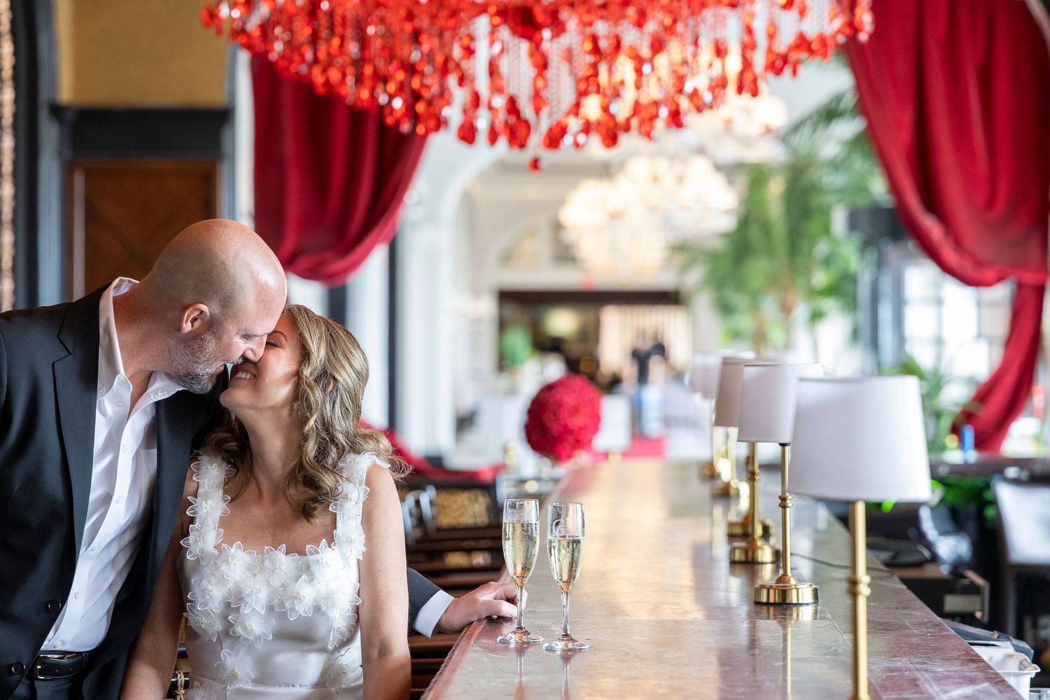 A couple in formal wedding attire sits at an elegant table with red curtains and crystal chandeliers in the background.