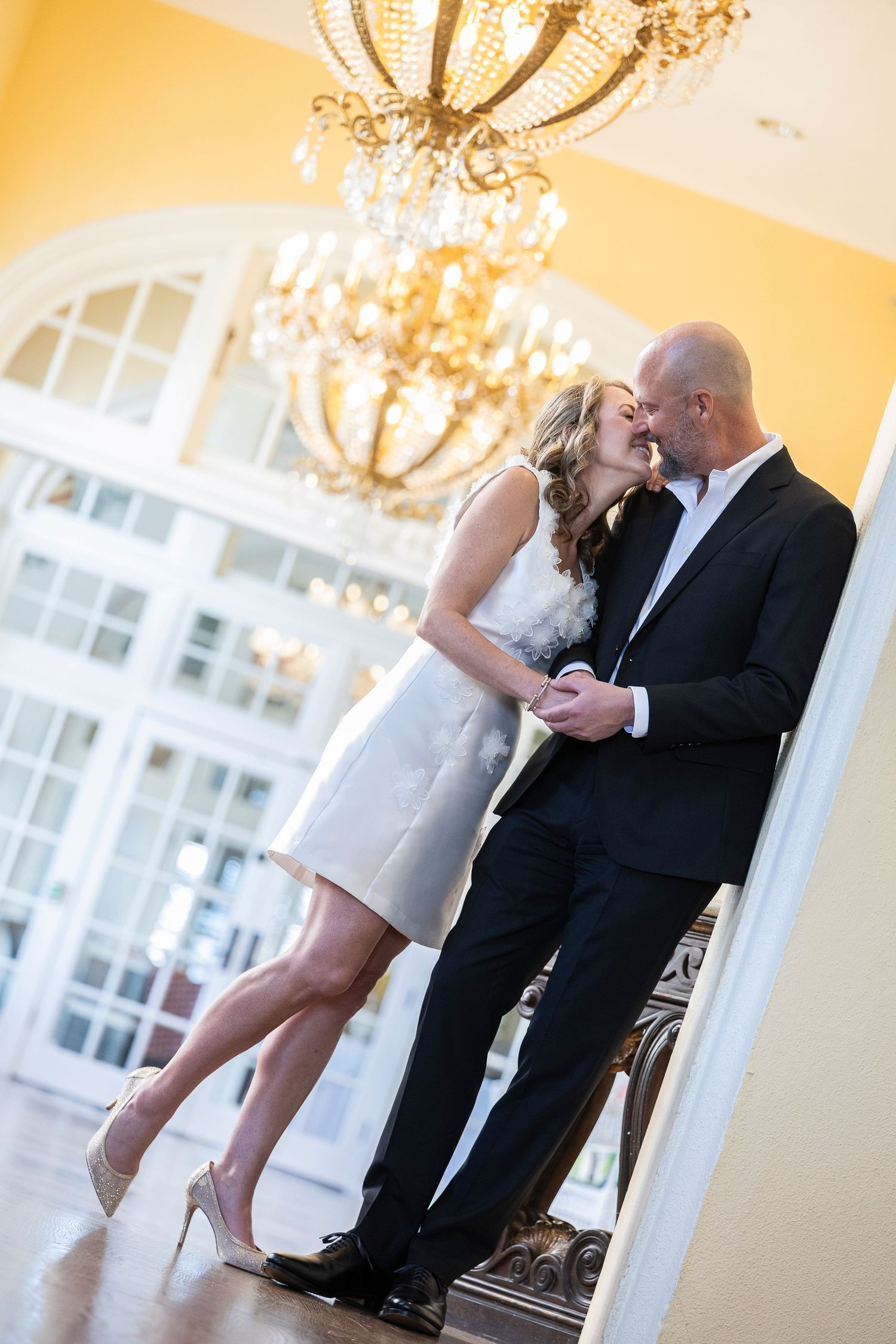 A romantic moment shared under an elegant crystal chandelier in a bright interior with white arched windows.