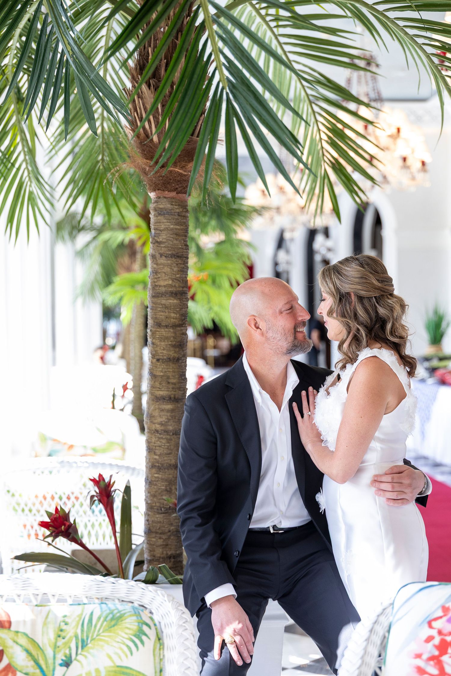 Couple sharing an intimate moment under tropical palm trees at an outdoor venue with white decor.