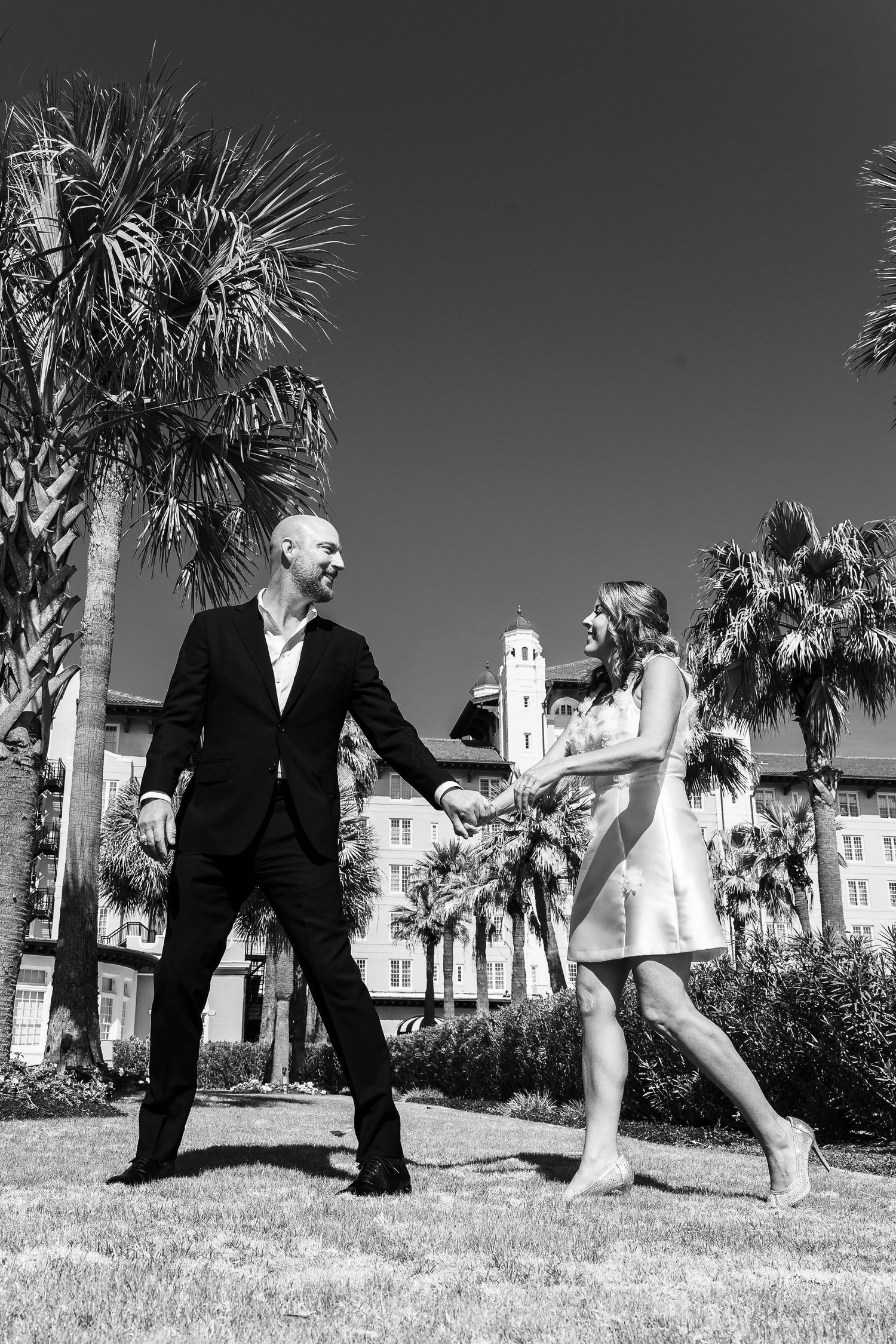 Black and white photo of two people playfully interacting on a lawn with palm trees and a historic building in background.