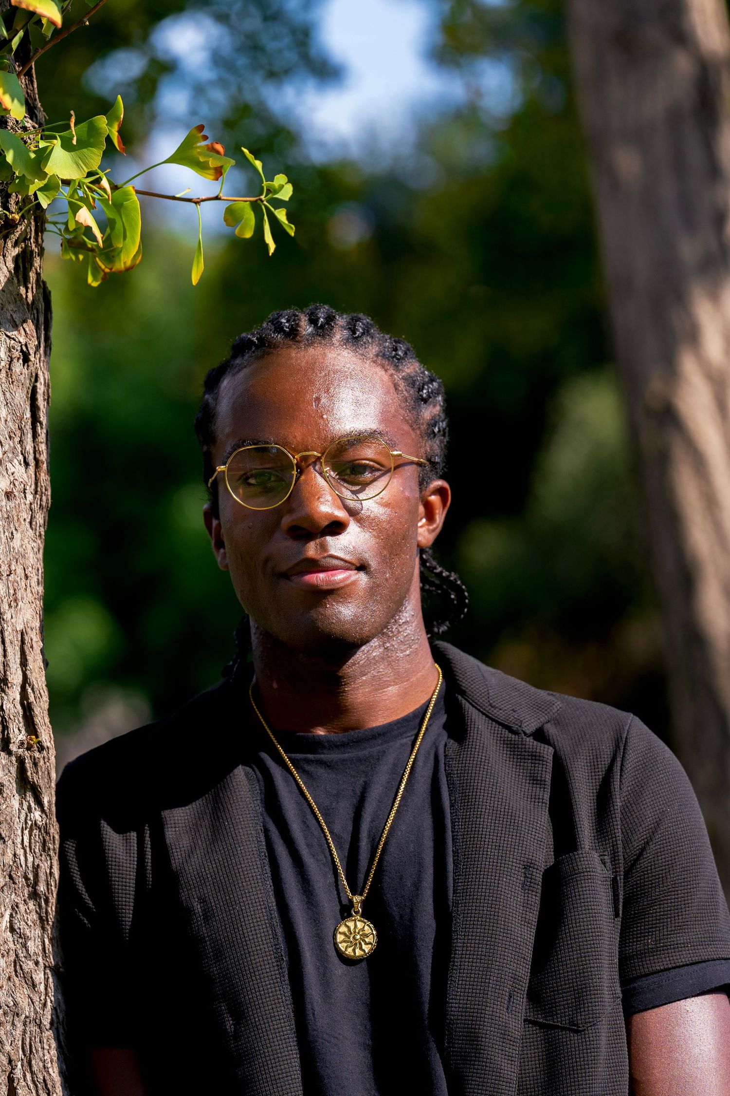 A portrait photo of Mwendwa Rwakatare leaning against a tree in a park. He has cornrows is wearing a black shirt and has a gold necklace on.