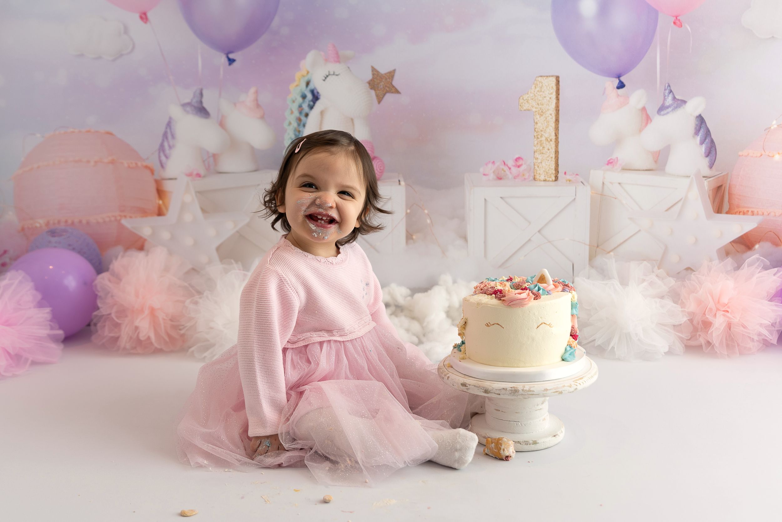 First birthday celebration setup with pink dress, white cake, purple balloons and tulle decorations against white backdrop.