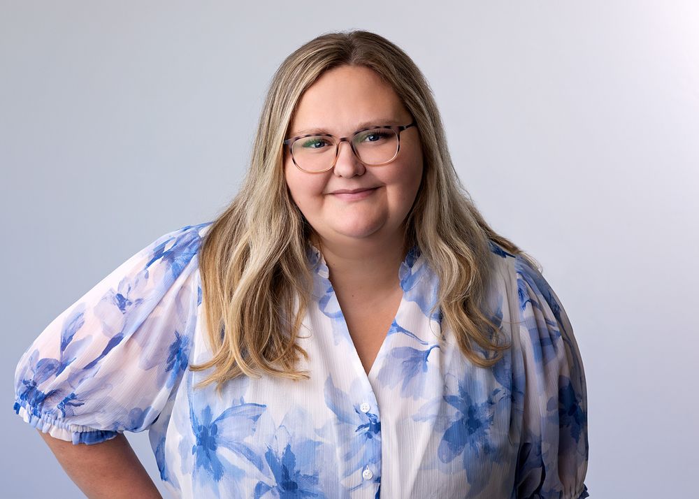 Woman with blonde hair and glasses smiling, wearing a blue floral blouse against a light gray background.