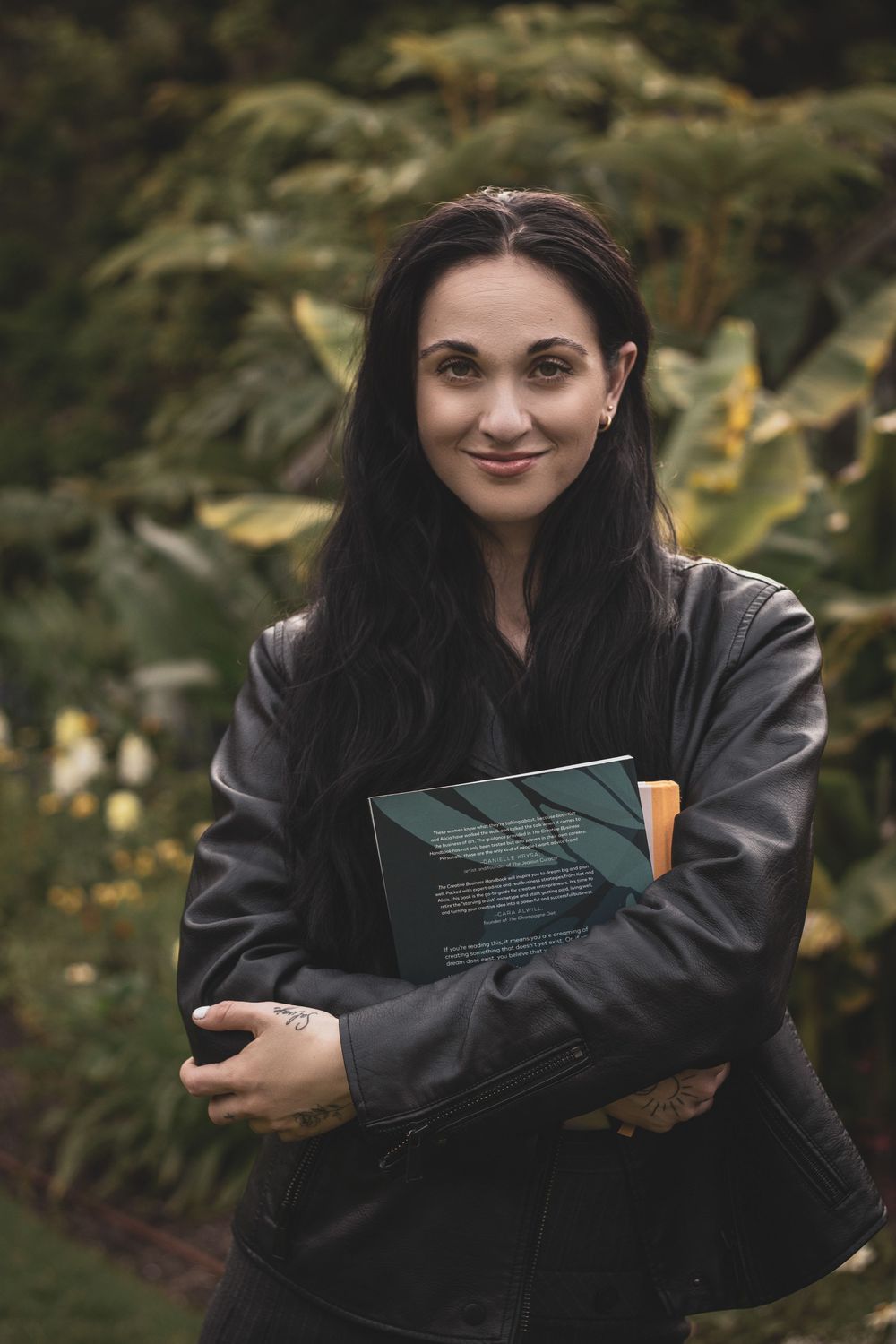 Person in black leather jacket holding green book while standing in outdoor garden setting with autumn foliage.